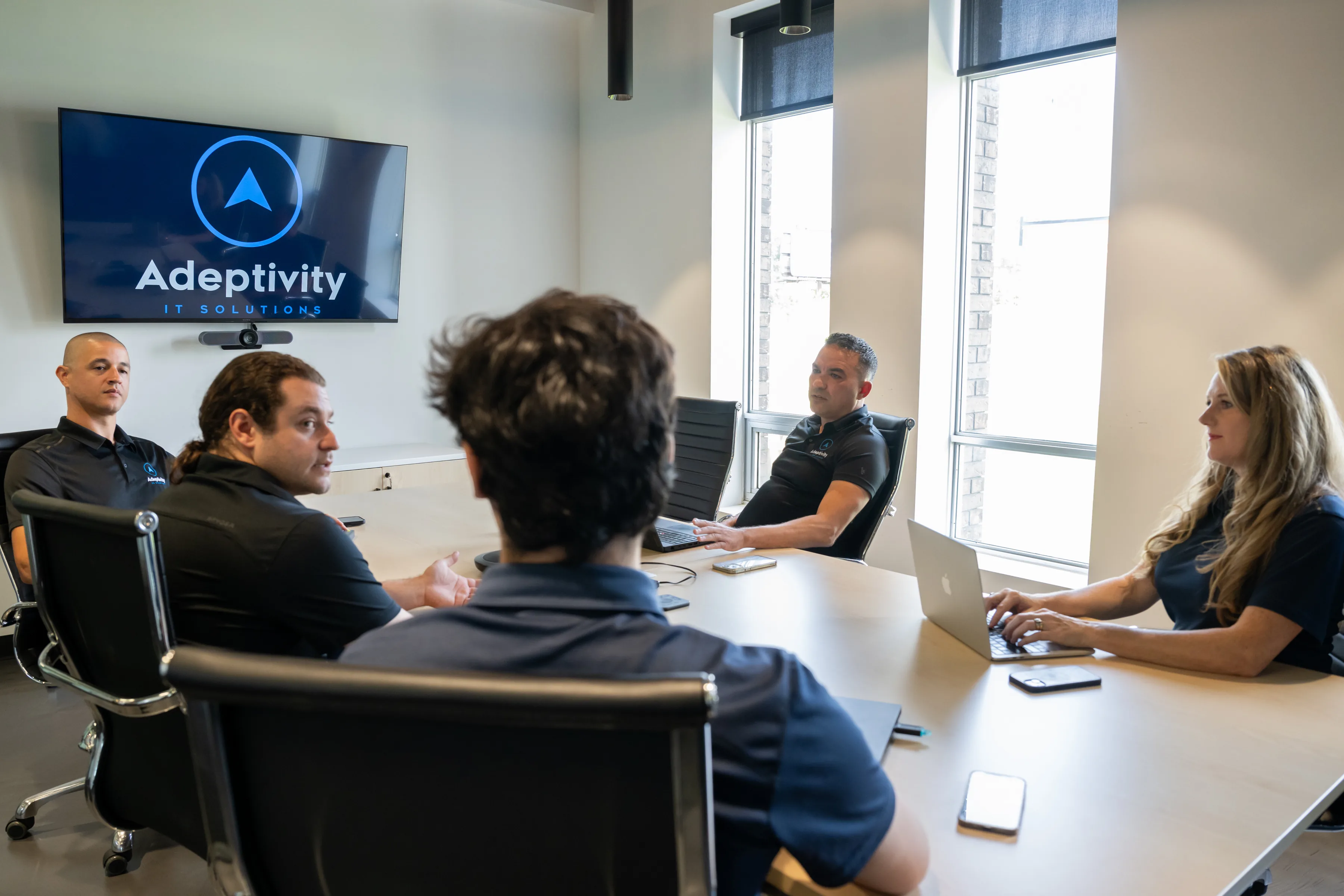 Five people in an office conference room having a meeting; three men and one woman facing each other around a light-colored table, one man is speaking, a wall-mounted screen displays the logo 'Adeptivity IT Solutions', and two large windows with roller shades provide natural light.