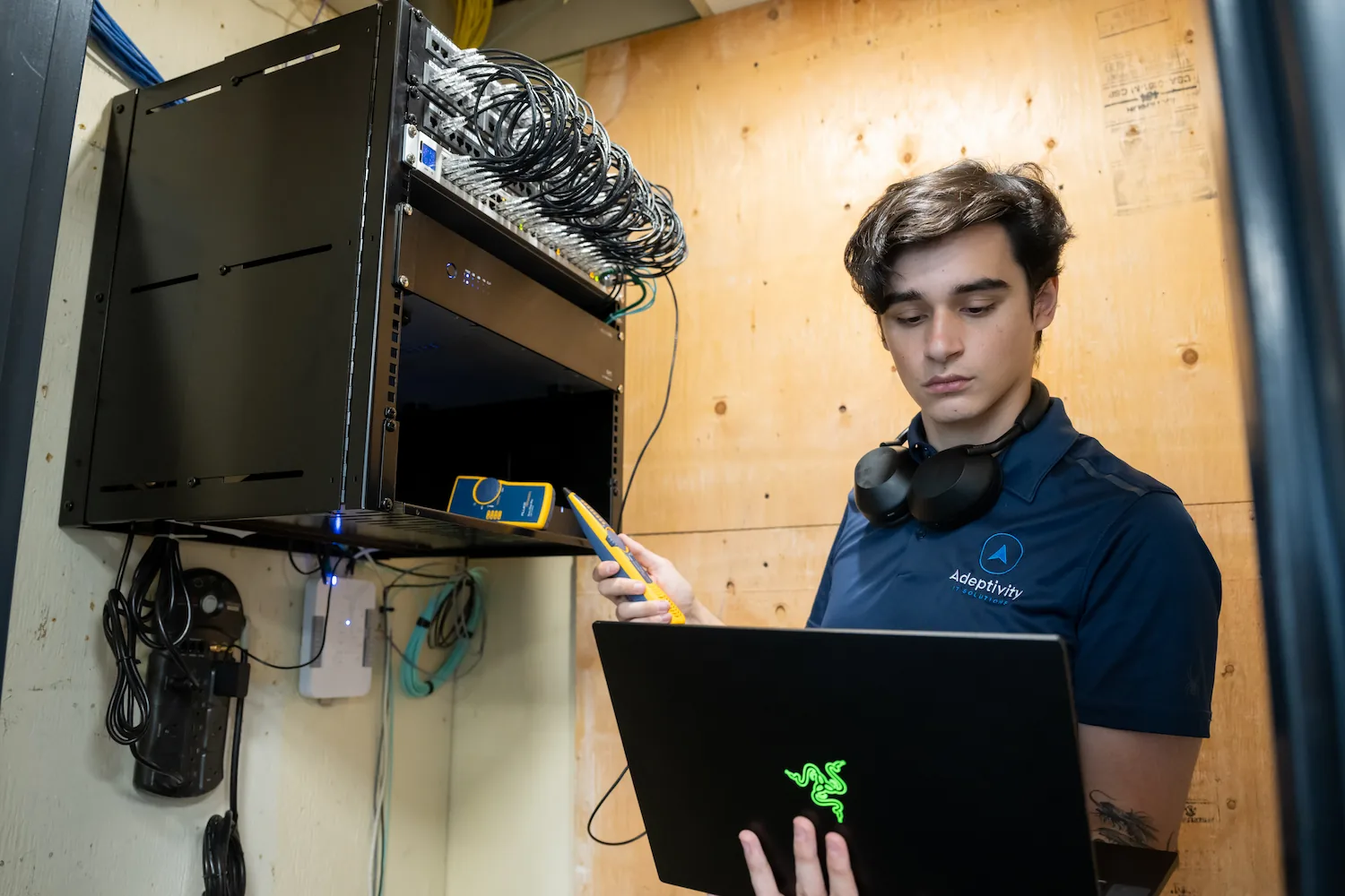 Technician wearing a navy Adeptivity Solutions polo and black headphones around his neck, holding a yellow network cable tester in one hand and a laptop in the other, working near a wall-mounted black server rack with network cables connected.