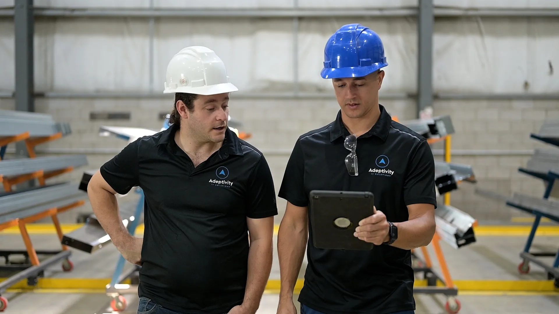 Two men wearing black Adeptivity IT Solutions shirts and safety helmets, one white and one blue, standing in an industrial warehouse; the man with the blue helmet is holding a tablet while the other looks on attentively.