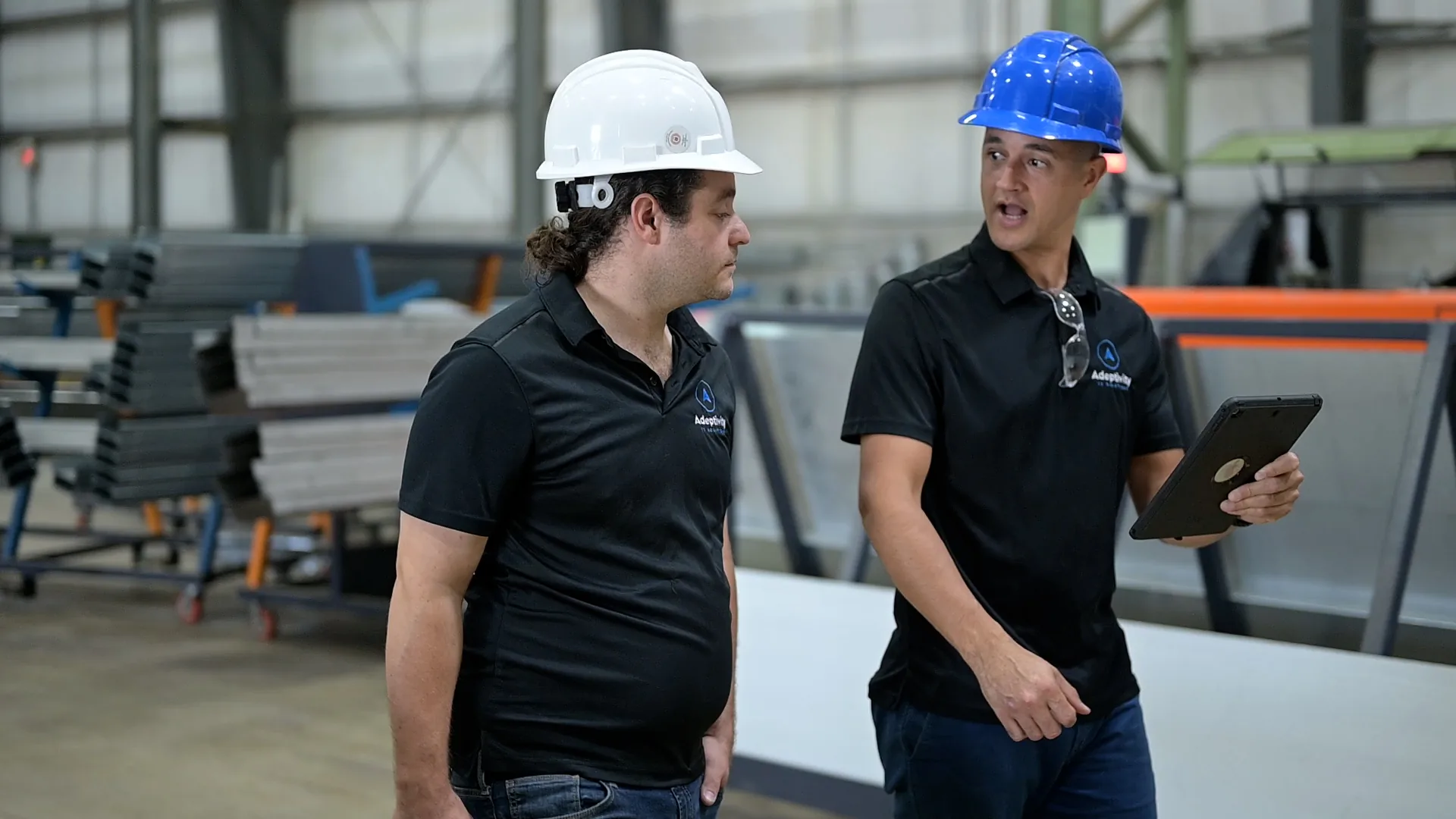 Two men wearing construction helmets and black polo shirts with a company logo are in a warehouse; one man holds a tablet and talks, while the other listens attentively.