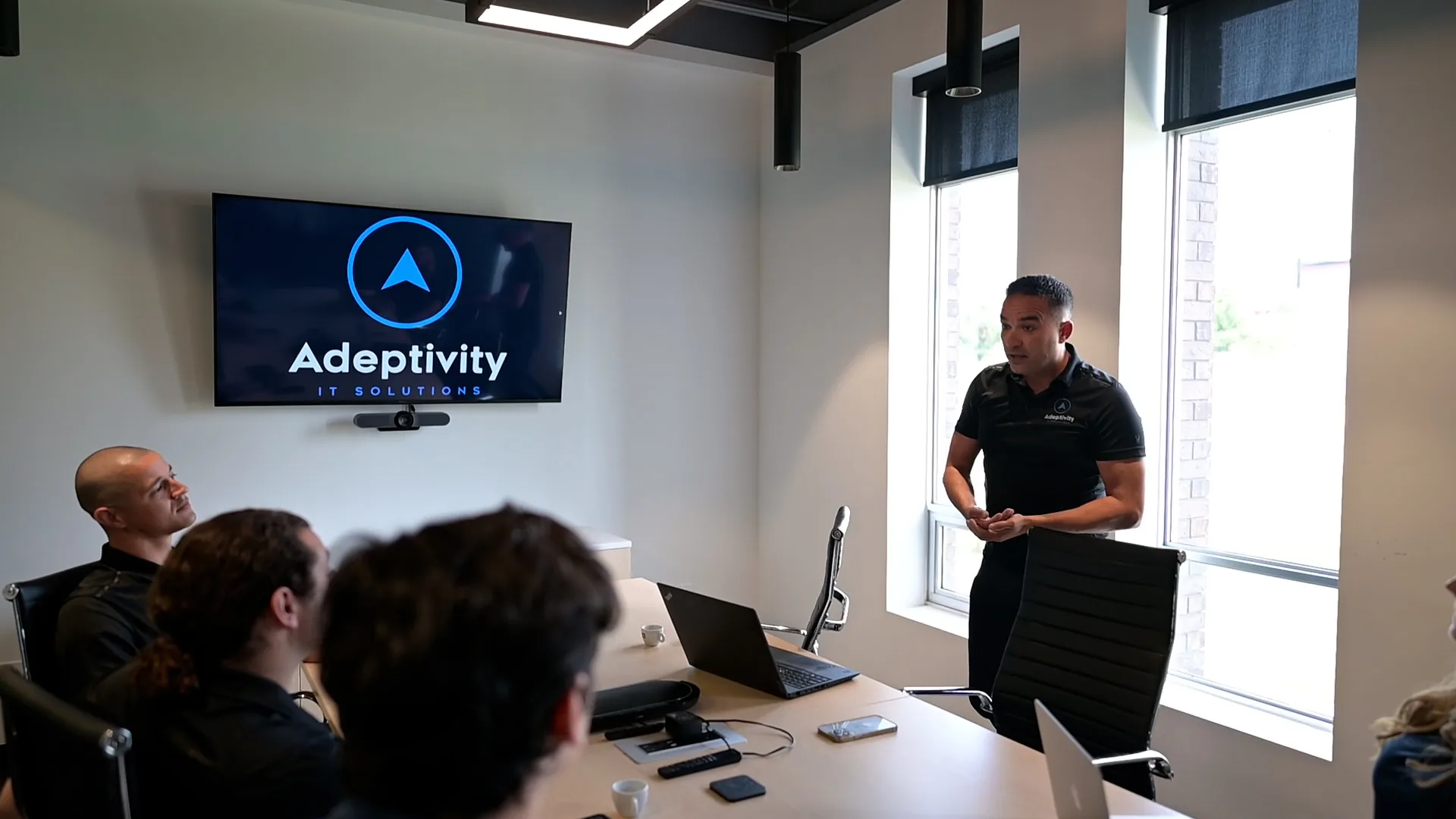 A man wearing a black Adeptivity IT Solutions polo shirt is standing and speaking in a meeting room to seated colleagues, with a wall-mounted screen displaying the Adeptivity IT Solutions logo behind him.