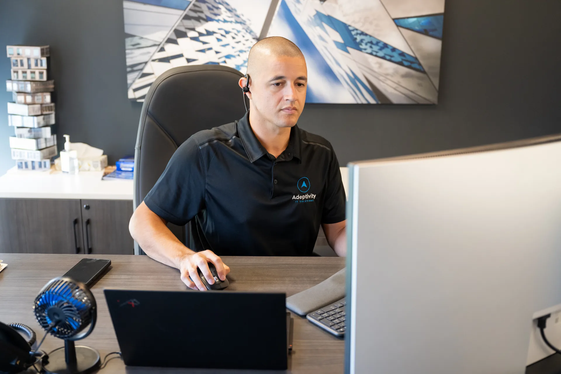 Man with a headset sitting at a desk using a computer mouse and working on a desktop computer, wearing a black polo shirt with Adeptivity IT Solutions logo.