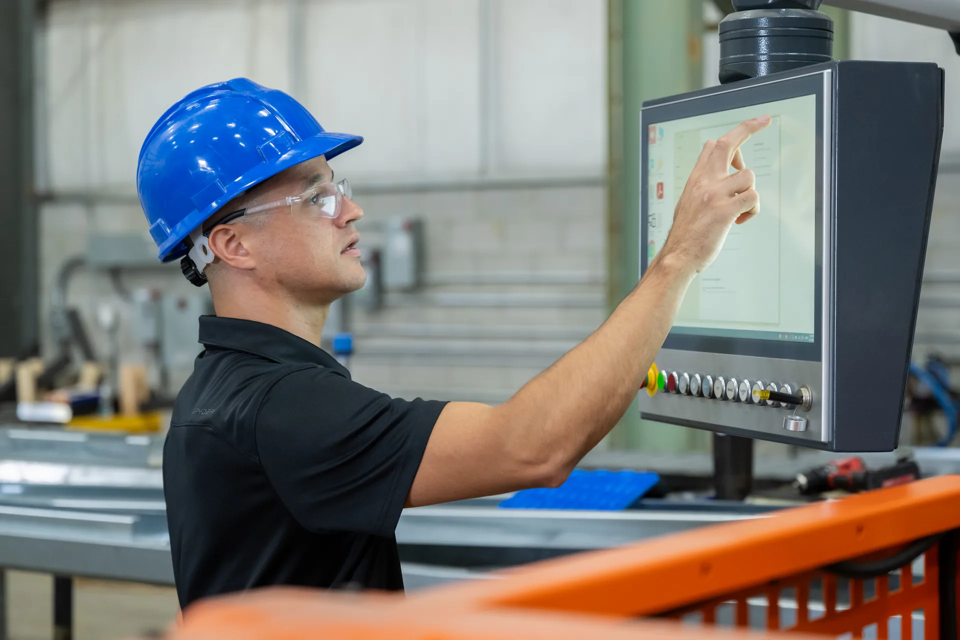 Male factory worker wearing a blue hard hat and safety glasses operating a touchscreen control panel in an industrial setting.