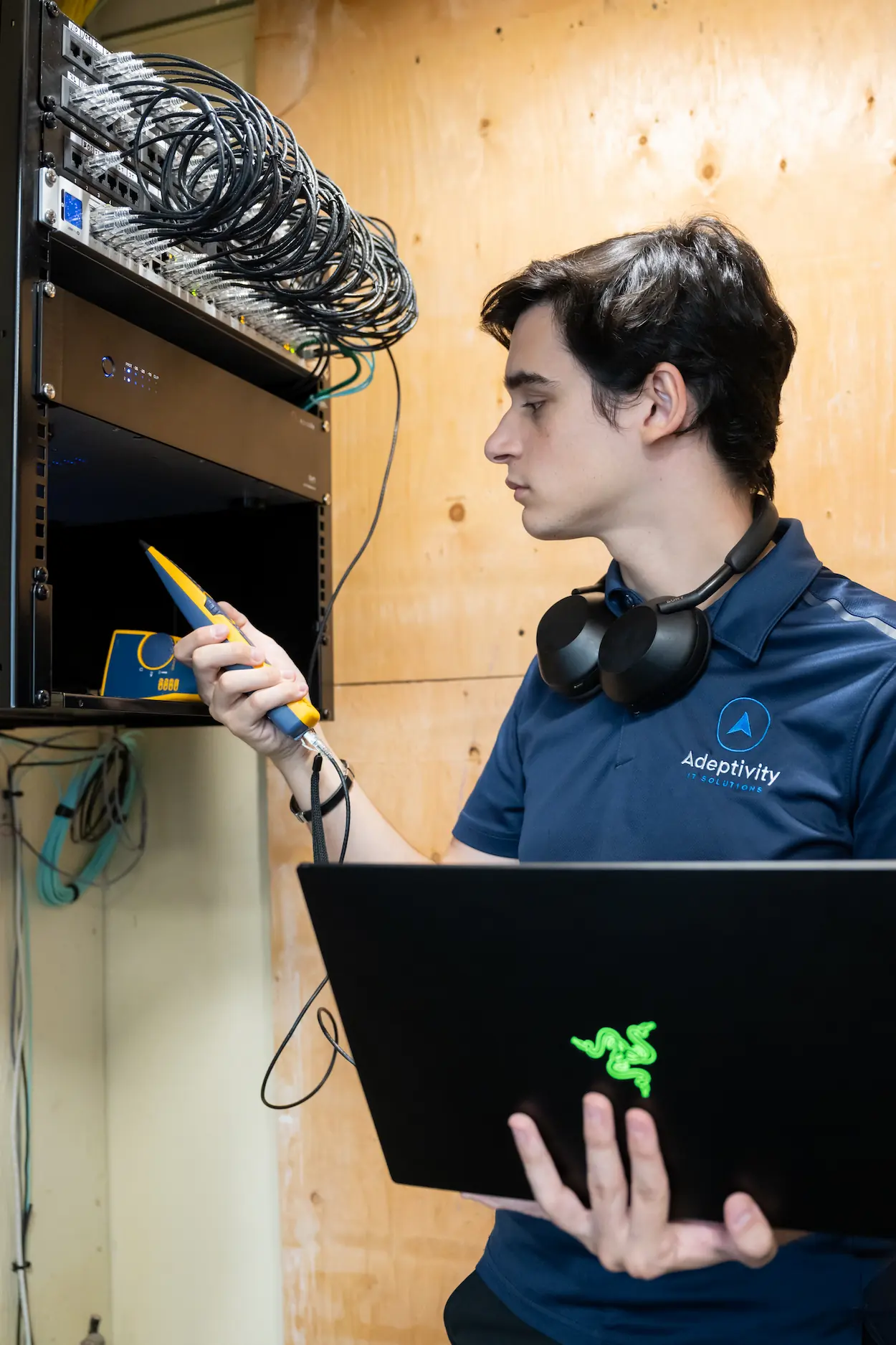 Technician in a blue Adeptivity IT Solutions polo shirt using a laptop and a handheld device to check a rack of network cables and switches mounted on a wooden wall.
