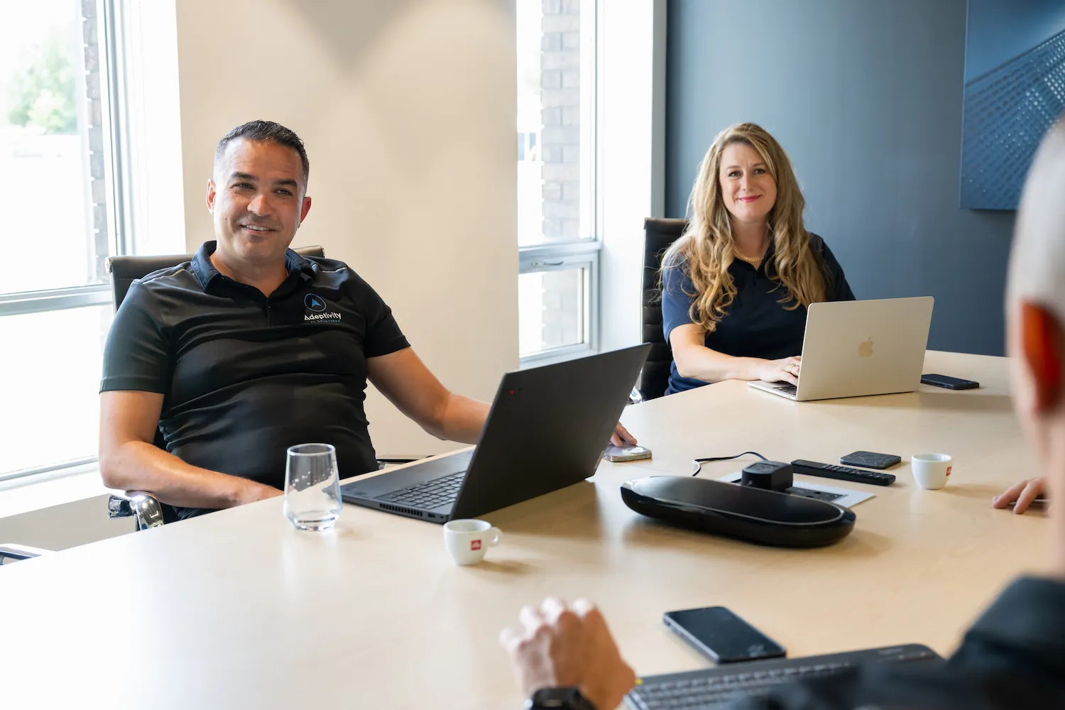 Two people sitting at a conference table with laptops in front of them, engaging in a meeting with a third person whose blurred hand and head appear in the foreground.