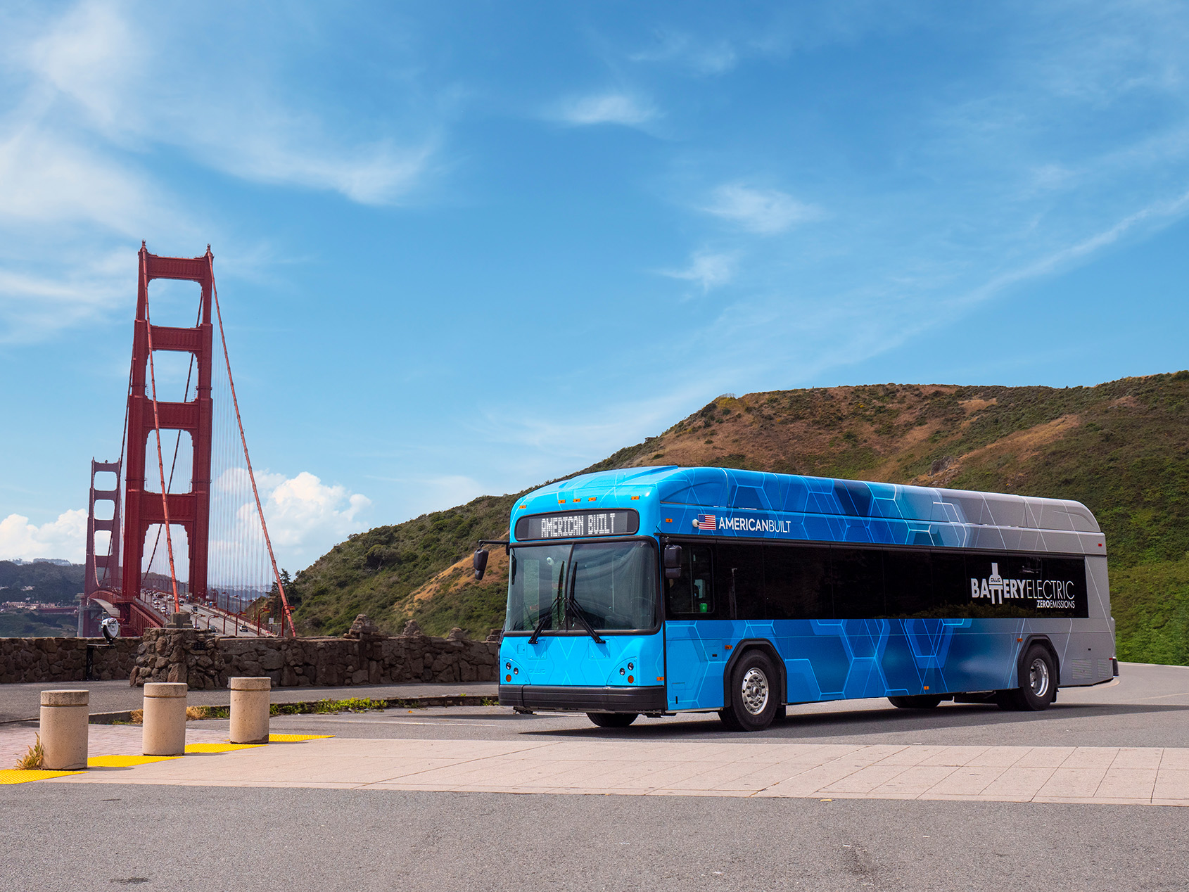 Blue and gray battery electric bus with American flag and 'American Built' text parked near the Golden Gate Bridge on a clear day with hills in the background.