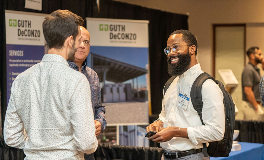 Three men engaged in conversation at a professional event booth with Guth DeConzo banners in the background; one man with a beard and glasses smiles while holding a phone and wearing a backpack.