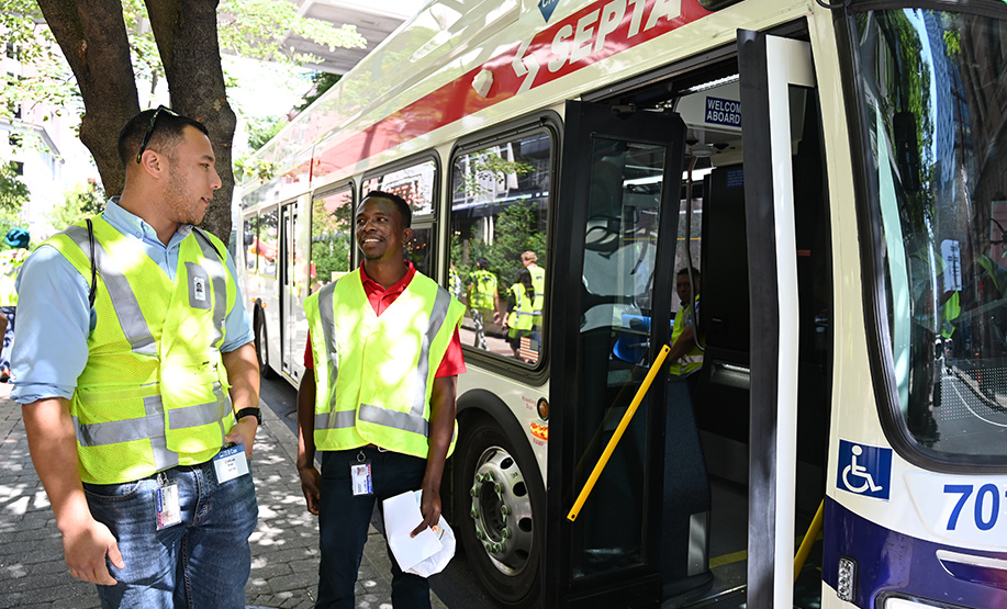 Two transit workers wearing yellow safety vests standing next to an open door of a SEPTA bus on a tree-lined street with reflections on the bus windows.