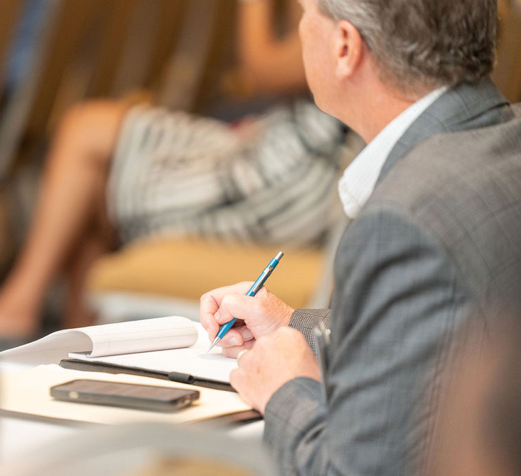 Man in a gray suit taking notes with a blue pen on a notepad, a smartphone lies on the table beside him, with a blurred background of another person sitting.