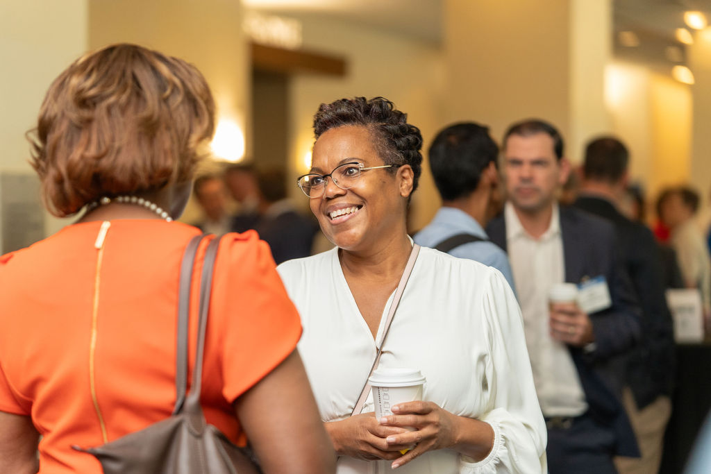 Smiling woman with short curly hair and glasses holding a coffee cup, talking to another woman in an orange top at a busy indoor event.