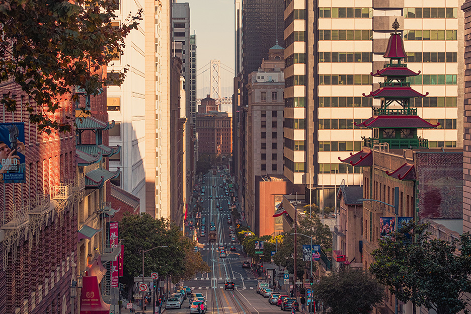 Urban street in San Francisco lined with tall buildings on both sides, featuring a pagoda-style roof on the right, cars parked along the street, and the Bay Bridge visible in the background.