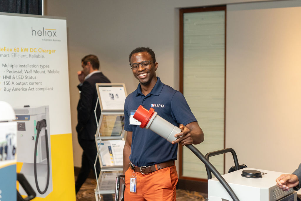 Smiling man in navy blue SEPTA polo and orange pants holding a large plug connected to a charging station at an indoor event with a Heliox 60 kW DC Charger banner in the background.