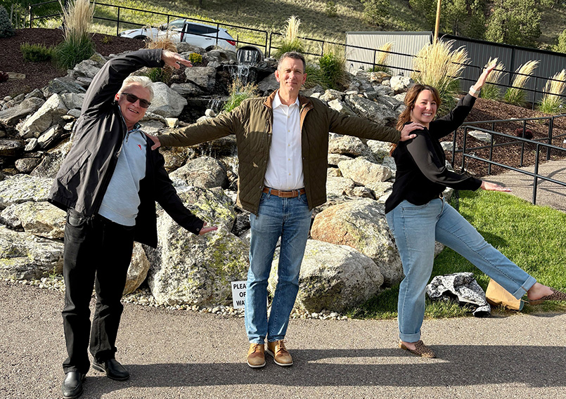 Three people standing outdoors on pavement in front of rocks and plants, posing to form the letters H, I, and ! with their arms and bodies, smiling.