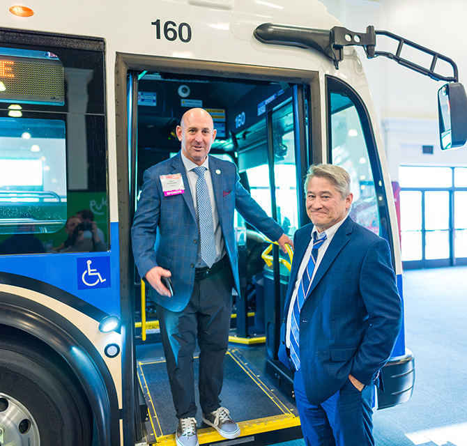Two men in business suits standing by the open door of a city bus numbered 160, with one man inside the bus and the other standing outside, inside a well-lit indoor space.
