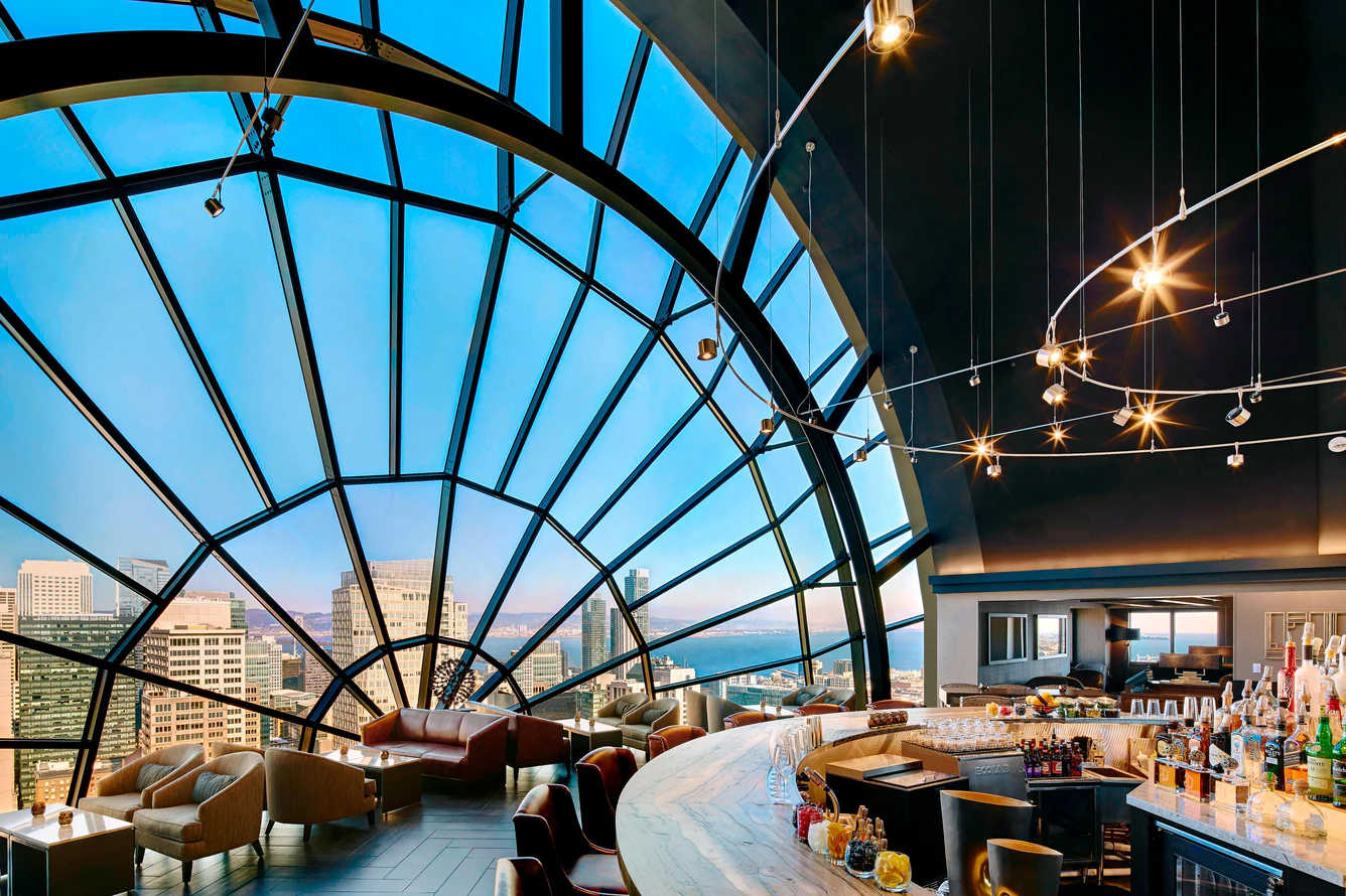 Modern hotel bar with a large curved window offering panoramic city and waterfront views, featuring comfortable seating and a well-stocked marble countertop bar under contemporary hanging lights.