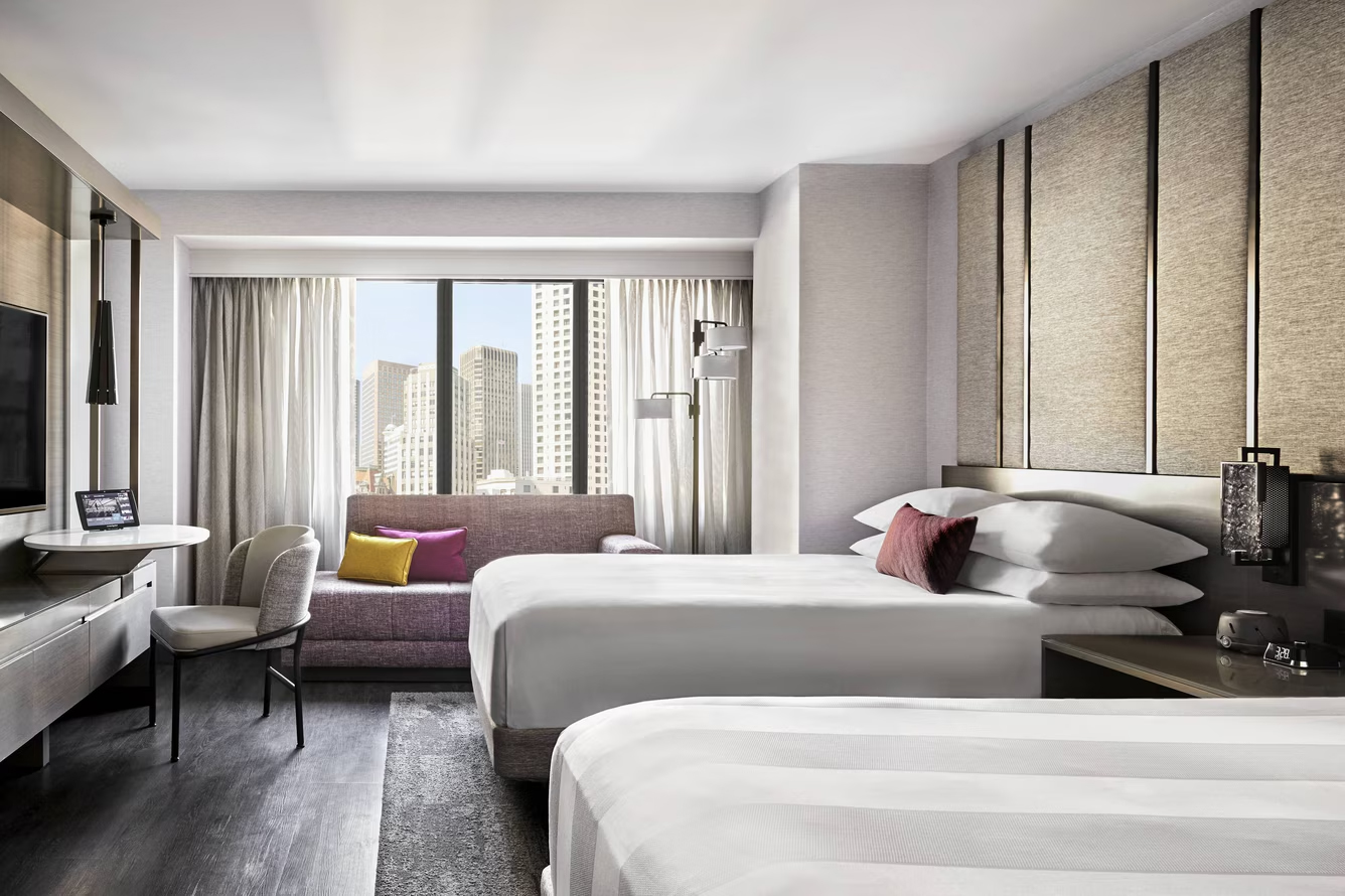 Modern hotel room with two white beds, a purple sofa with colorful pillows, a desk and chair near a large window showing city skyscrapers, and neutral-toned walls with vertical dark accents.