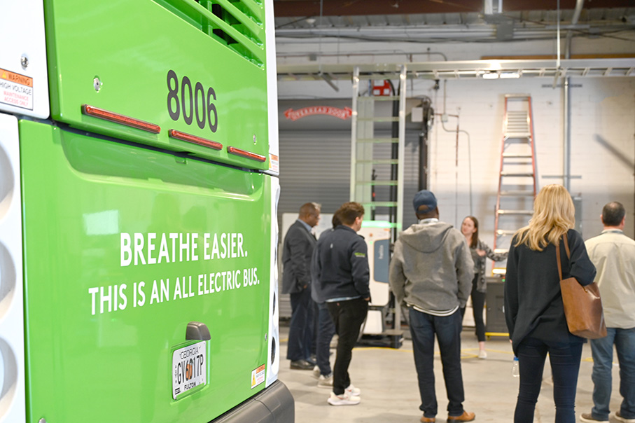 Green electric bus with text BREATHE EASIER. THIS IS AN ALL ELECTRIC BUS on its rear left side in a garage with a group of people standing and talking in the background.