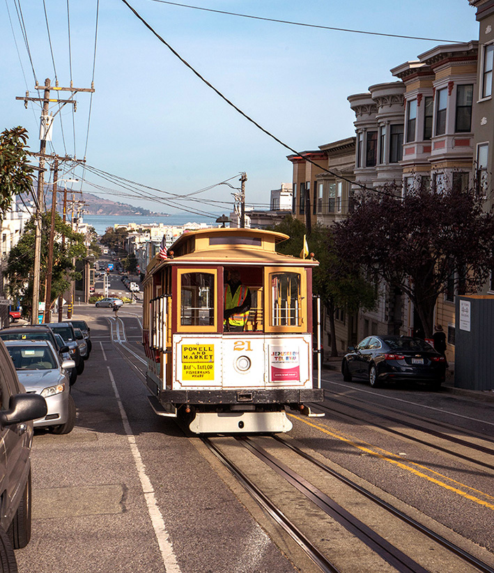 A vintage San Francisco cable car numbered 21 traveling uphill on a street lined with parked cars and multi-story houses, with a view of water and hills in the background.