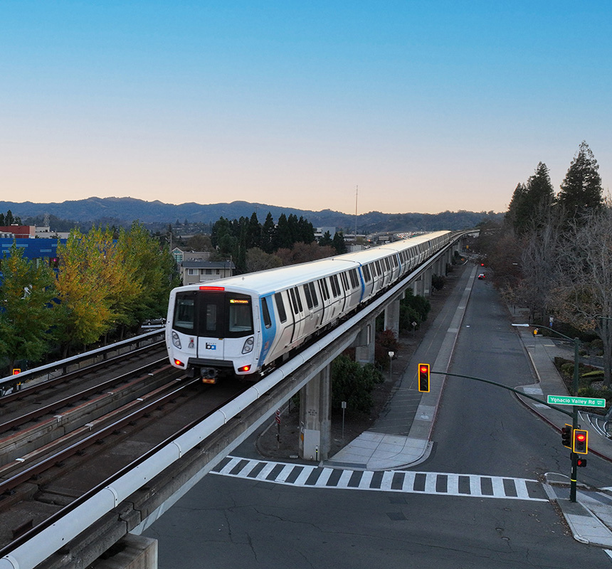 A BART train travels on an elevated track over a road intersection at sunset, with red traffic lights and trees on both sides, and mountains in the distance under a clear blue sky.