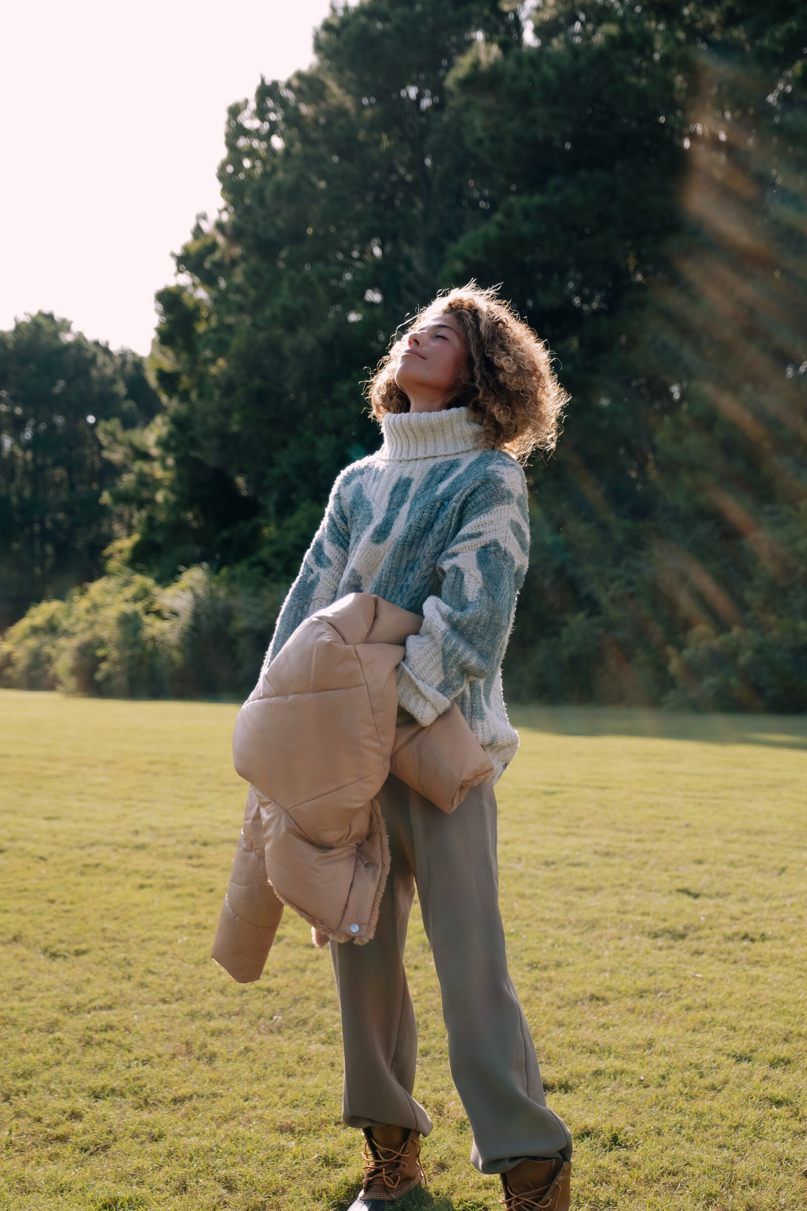 A woman standing outdoors in a sunlit green field, looking upward