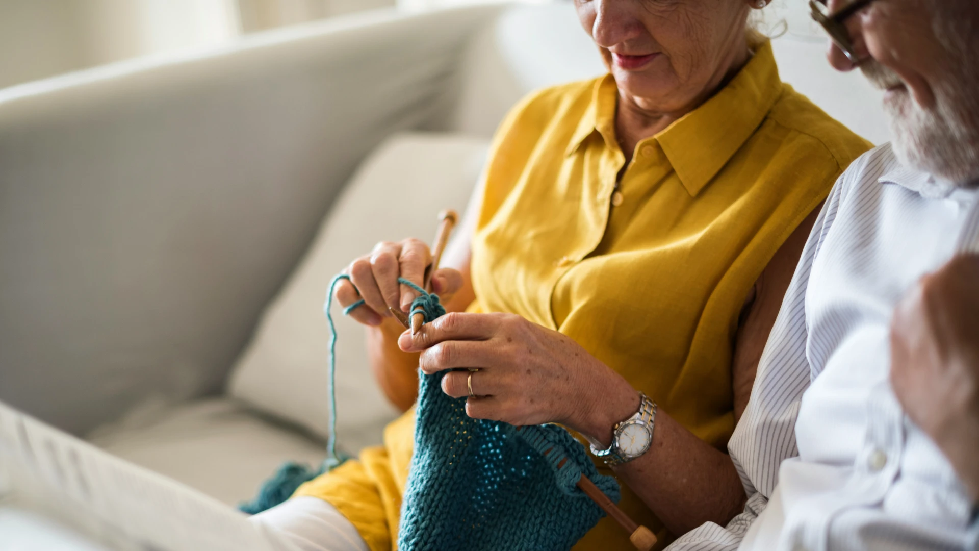 Elderly woman in a yellow sleeveless shirt knitting a blue piece with wooden needles while sitting next to an elderly man wearing glasses and a white striped shirt on a light-colored couch.