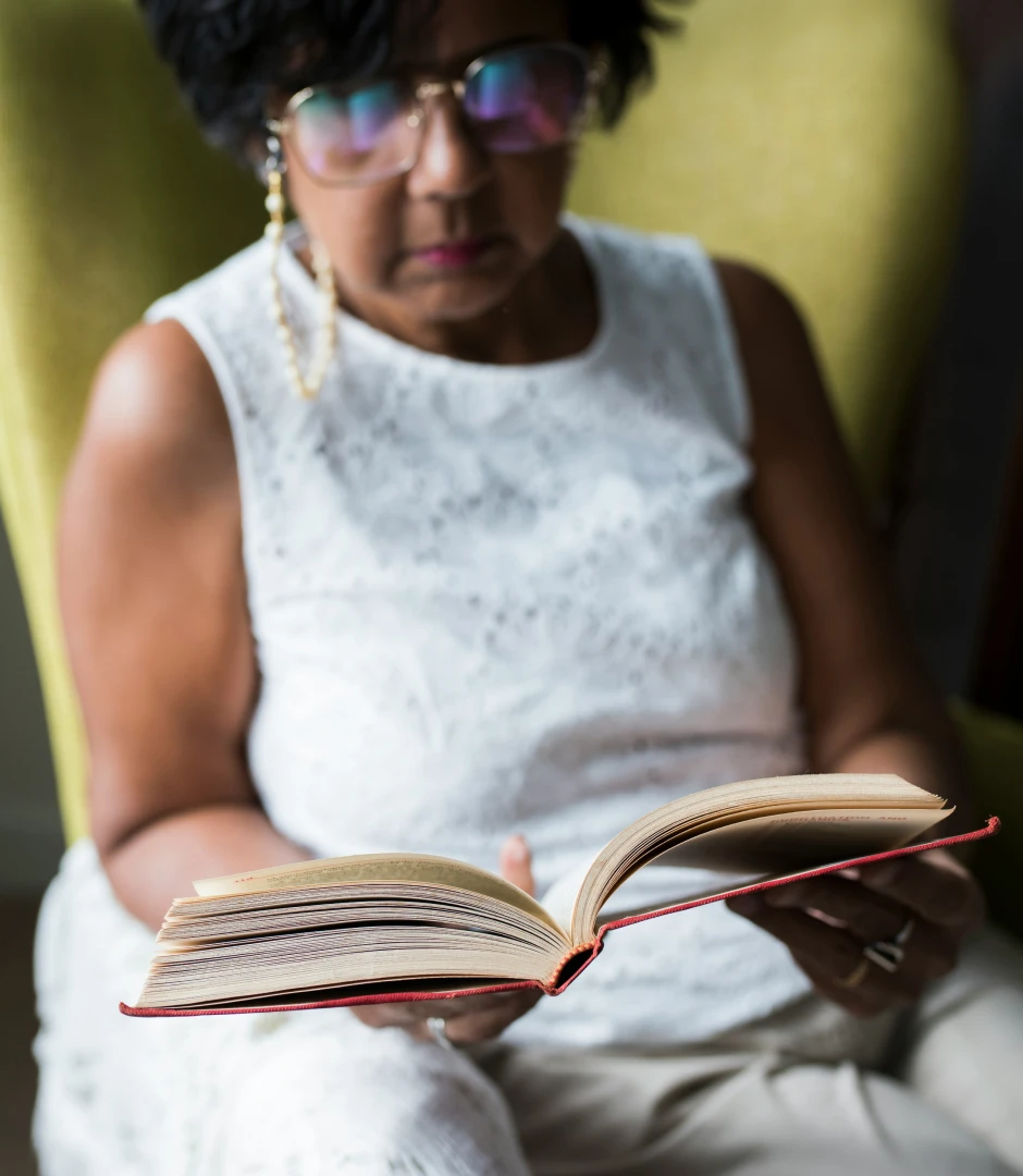 Middle-aged woman with glasses and a white lace sleeveless top sitting in a green chair, reading an open book held in both hands.