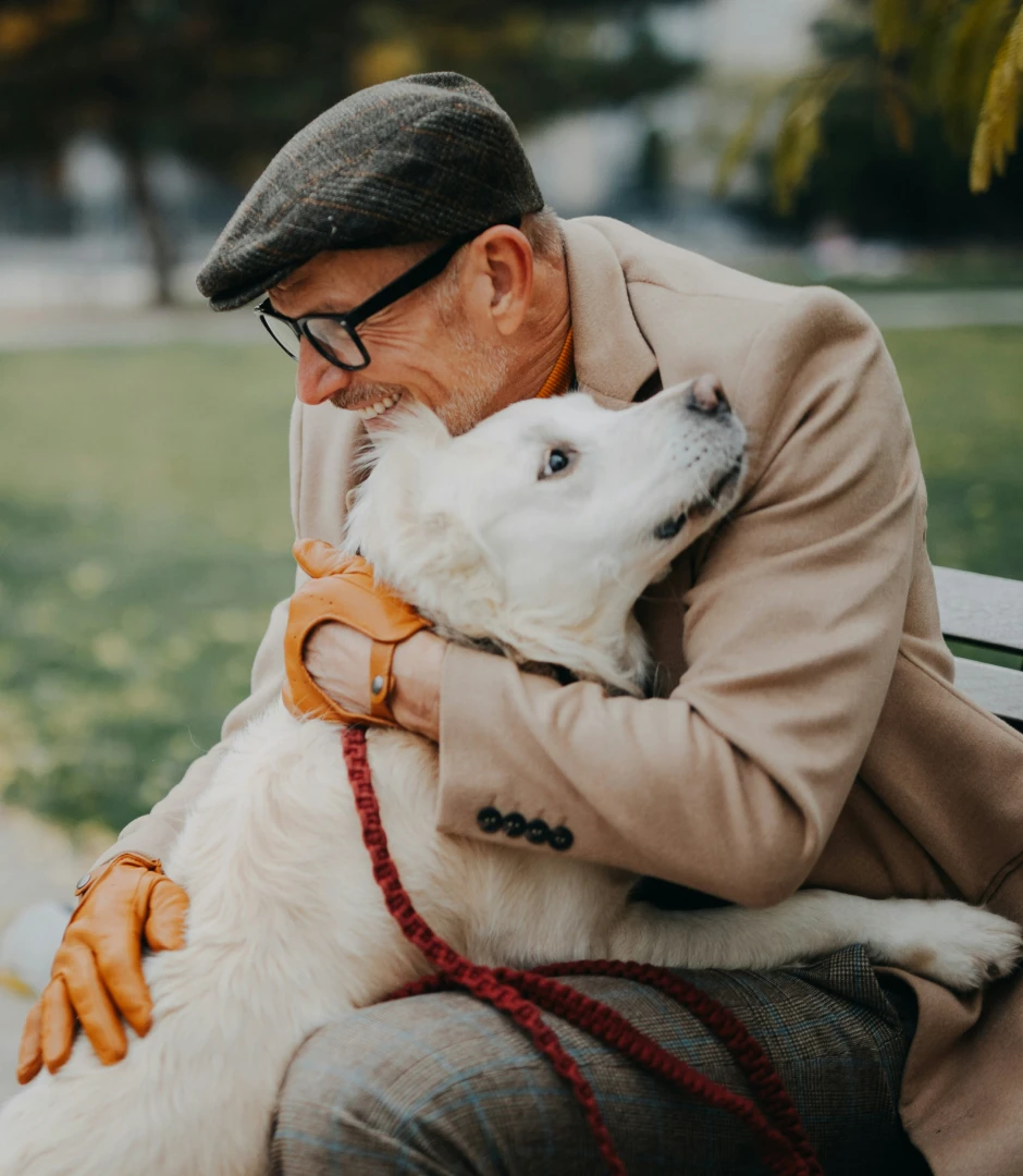 Smiling elderly man wearing a brown flat cap, glasses, beige coat, and orange gloves hugging a large white dog with a red leash while sitting on a park bench.