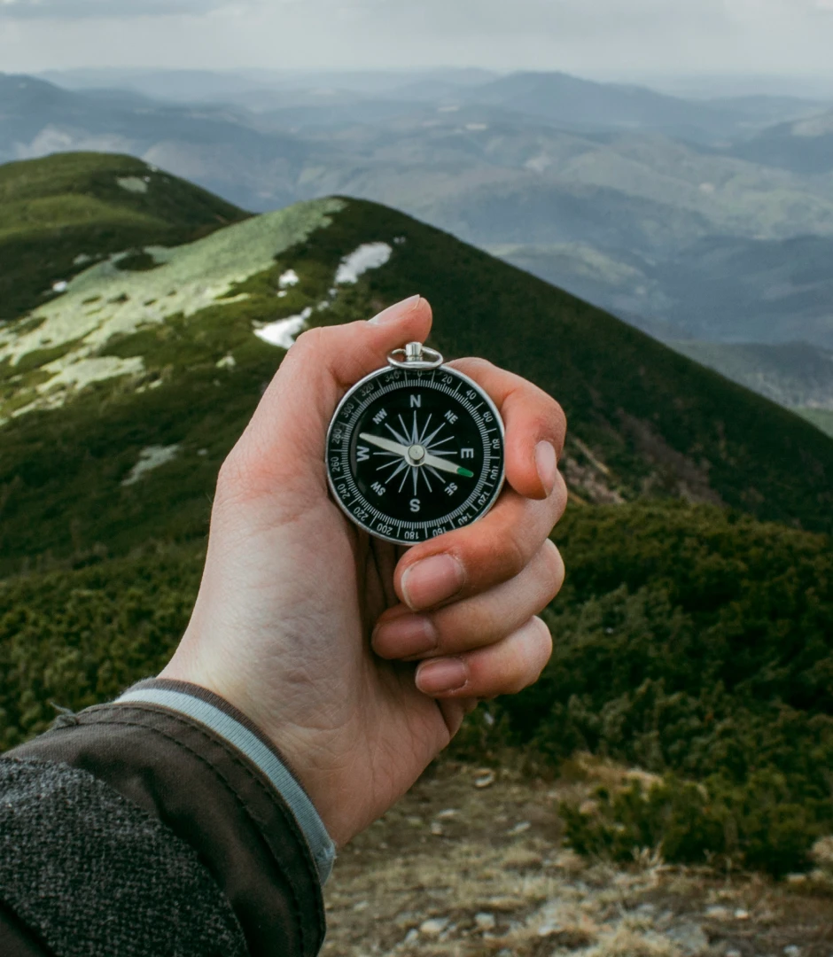 Close-up of a hand holding a black compass pointing north, with mountains and greenery in the background.