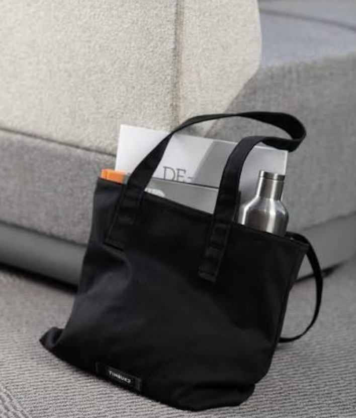 A black branded tote bag holding a magazine and an insulated steel bottle, styled as corporate merch