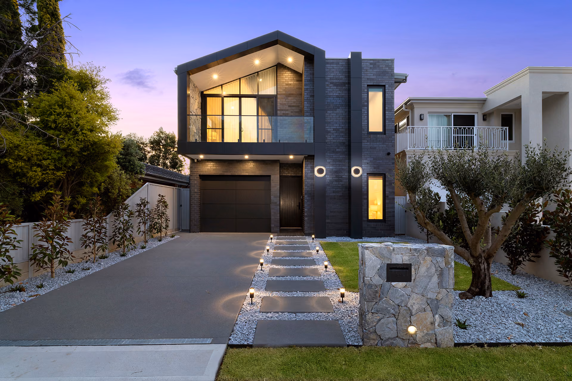 Modern two-story house with stone accents, illuminated pathway, and a manicured lawn at dusk.