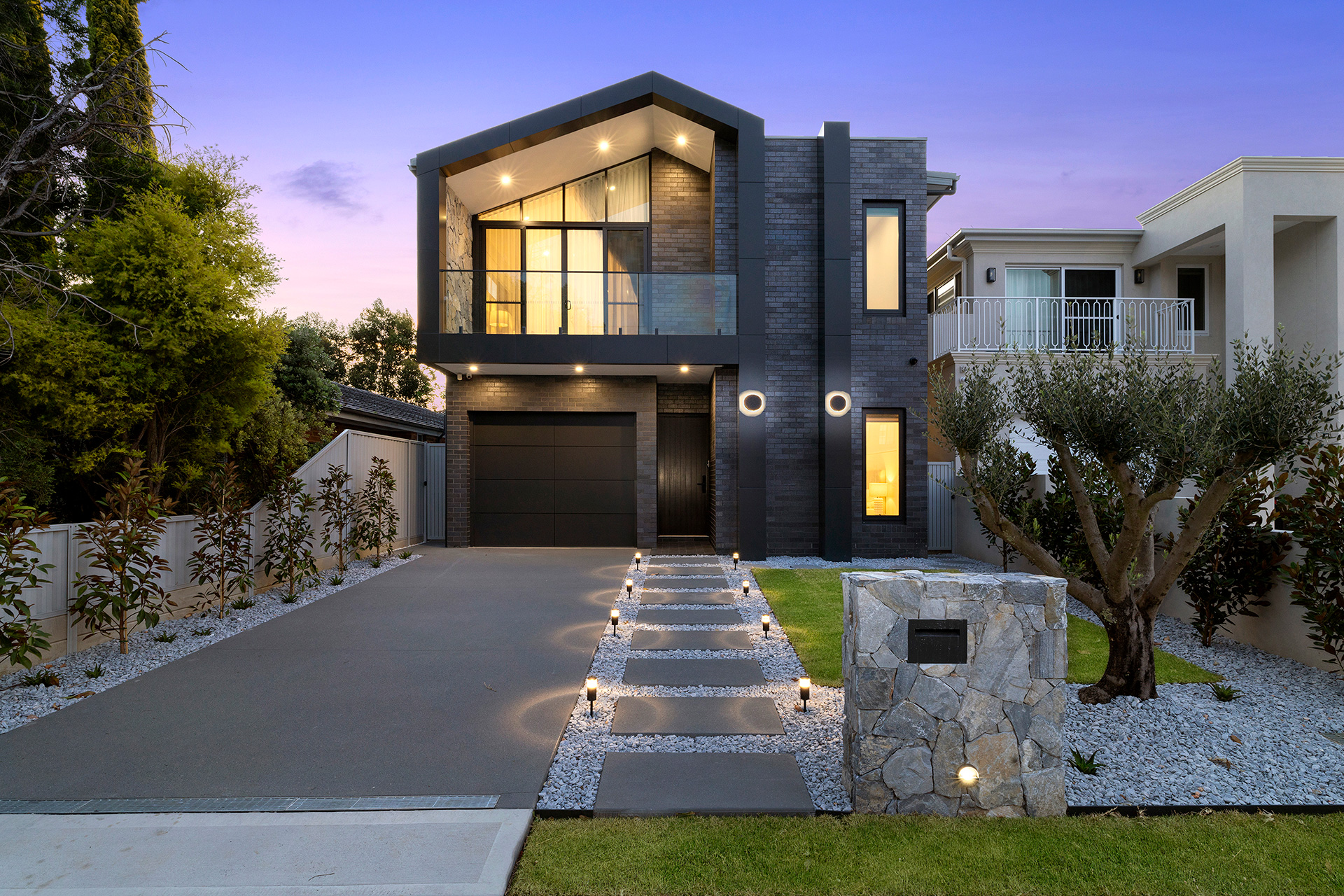 Modern two-story house with stone accents, illuminated pathway, and a manicured lawn at dusk.