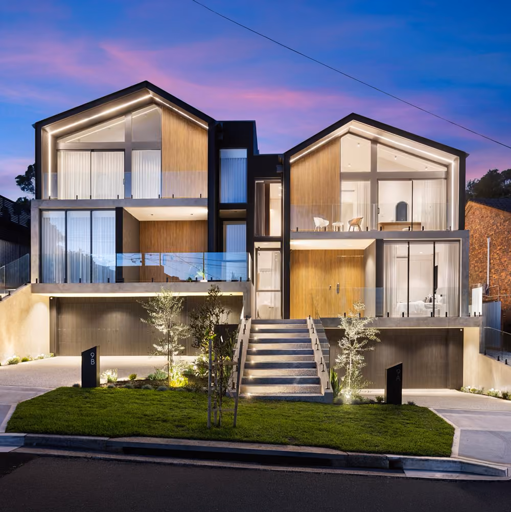 Modern twin house with illuminated balconies, stairway, garage doors, and landscaped front yard at dusk.