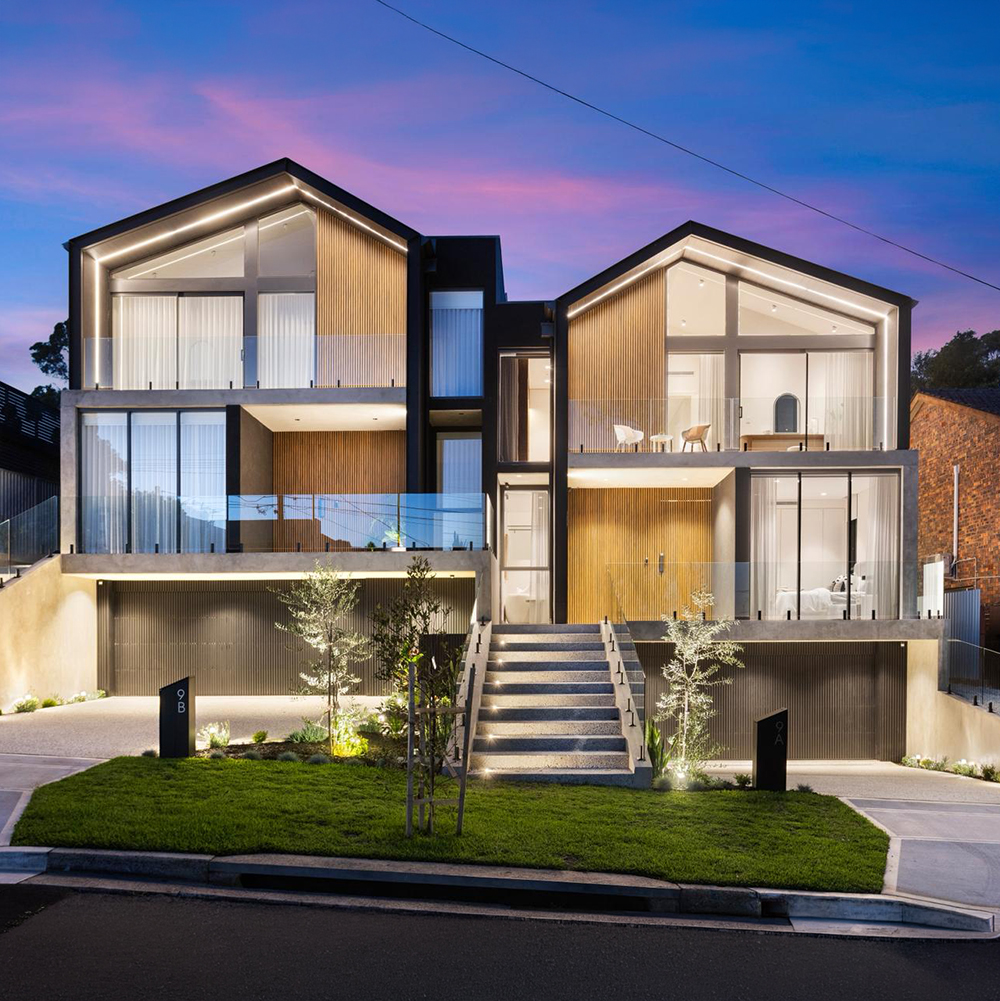 Modern twin house with illuminated balconies, stairway, garage doors, and landscaped front yard at dusk.