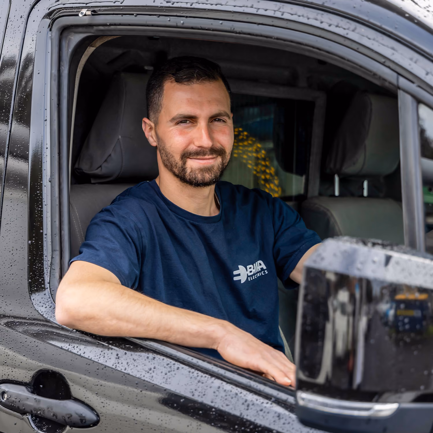 Man in a navy blue BMA Electrics shirt sitting in a black vehicle with rain droplets on the window.