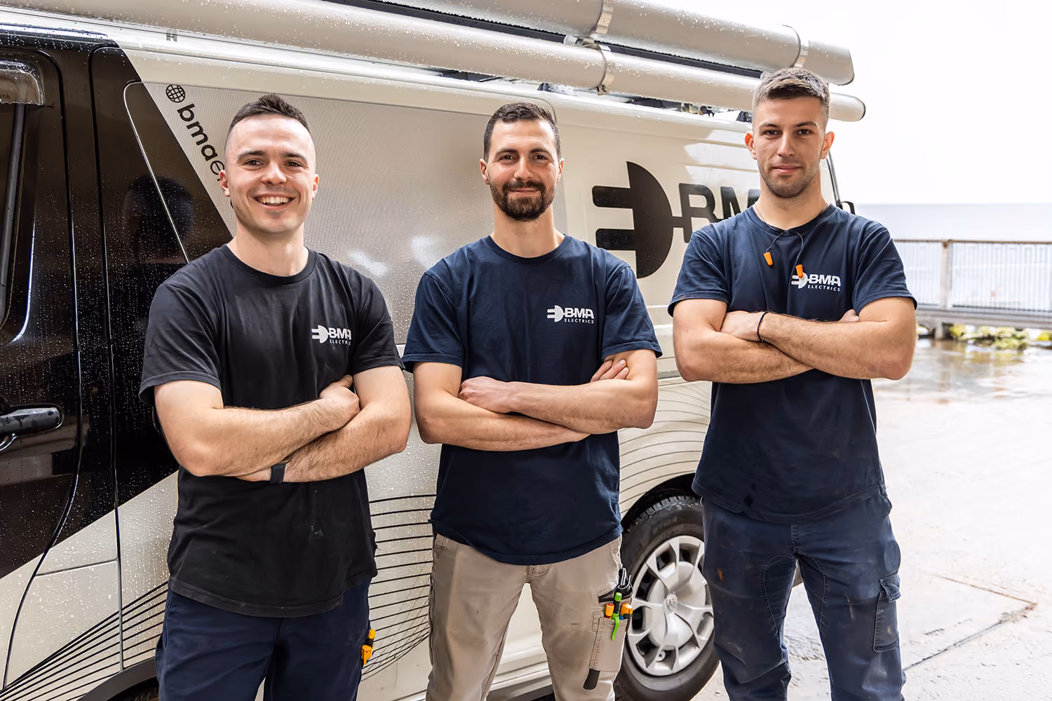 Three men wearing BMA Electrics shirts stand with arms crossed in front of a company van.