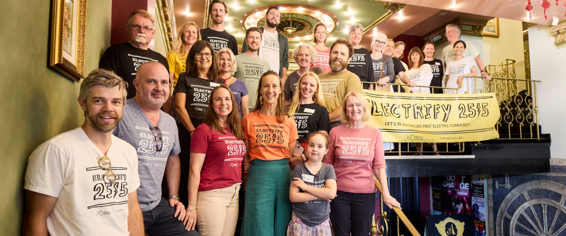 Group of diverse people of various ages standing on stairs and a landing inside a building, many wearing colorful t-shirts with 'Electrify 2515' printed on them, holding a large yellow banner reading 'Electrify 2515 Let's be Australia's first electric community.'