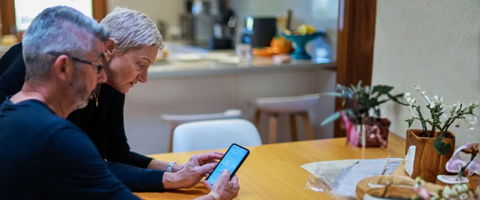An older man and woman sitting at a wooden table in a kitchen, looking at a smartphone screen together.