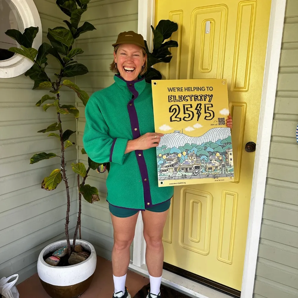 Smiling person standing in front of a yellow door holding a yellow sign that reads 'We're helping to electrify 2515'.