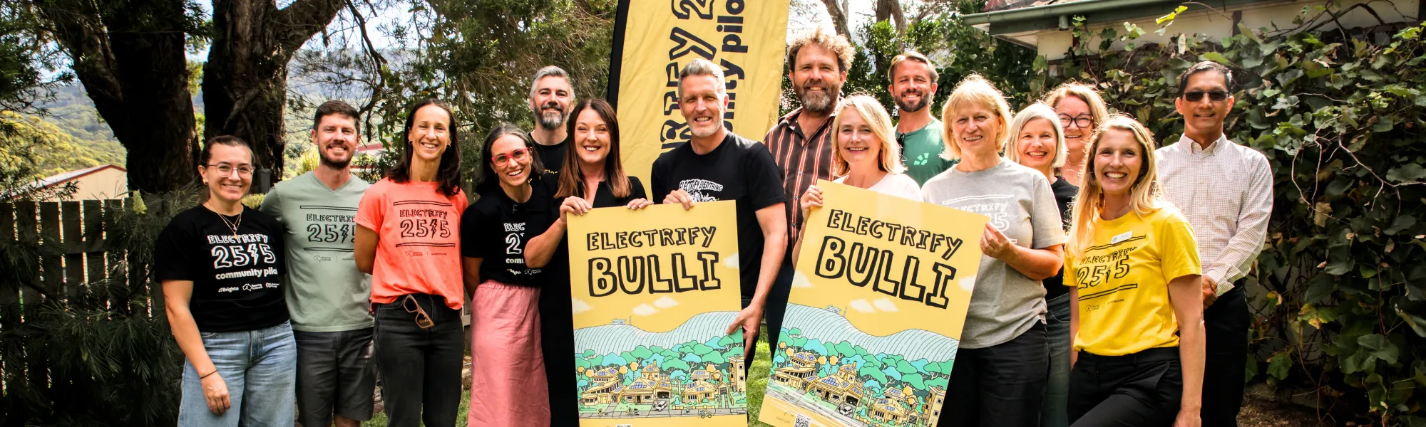 Group of diverse people standing outdoors in front of trees and a building, many wearing colorful Electrify 2515 community plan t-shirts, some holding two yellow posters with 'ELECTRIFY BULLI' text and illustrations of houses and hills.
