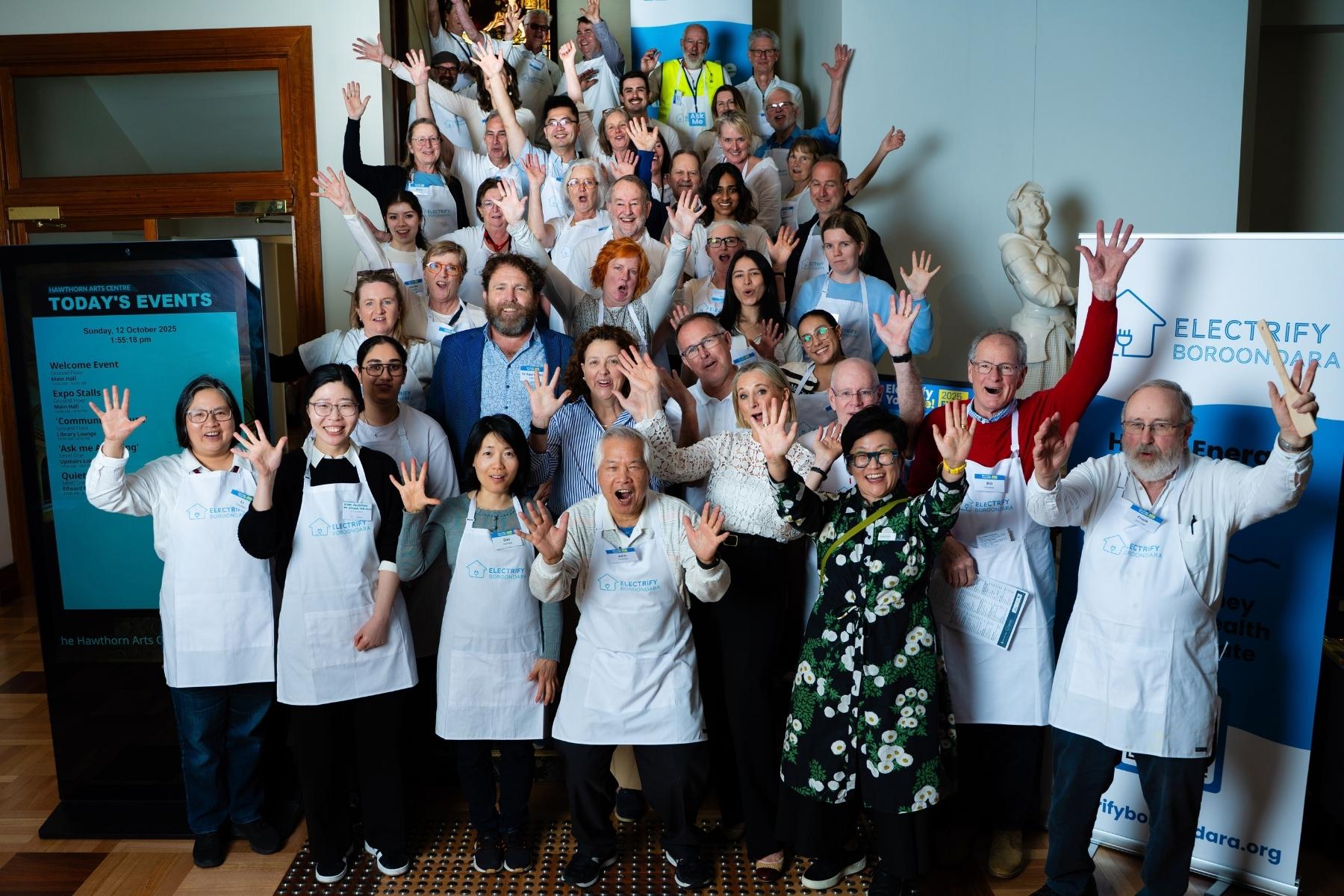 A large group of Electric Community volunteers in aprons cheering with their hands in the air at a community expo