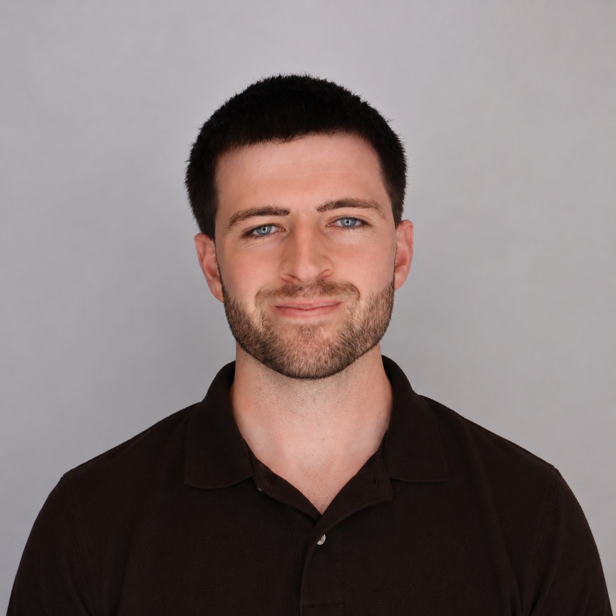 Portrait of a young man with short dark hair, blue eyes, and a beard, wearing a black collared shirt, smiling slightly against a plain gray background.