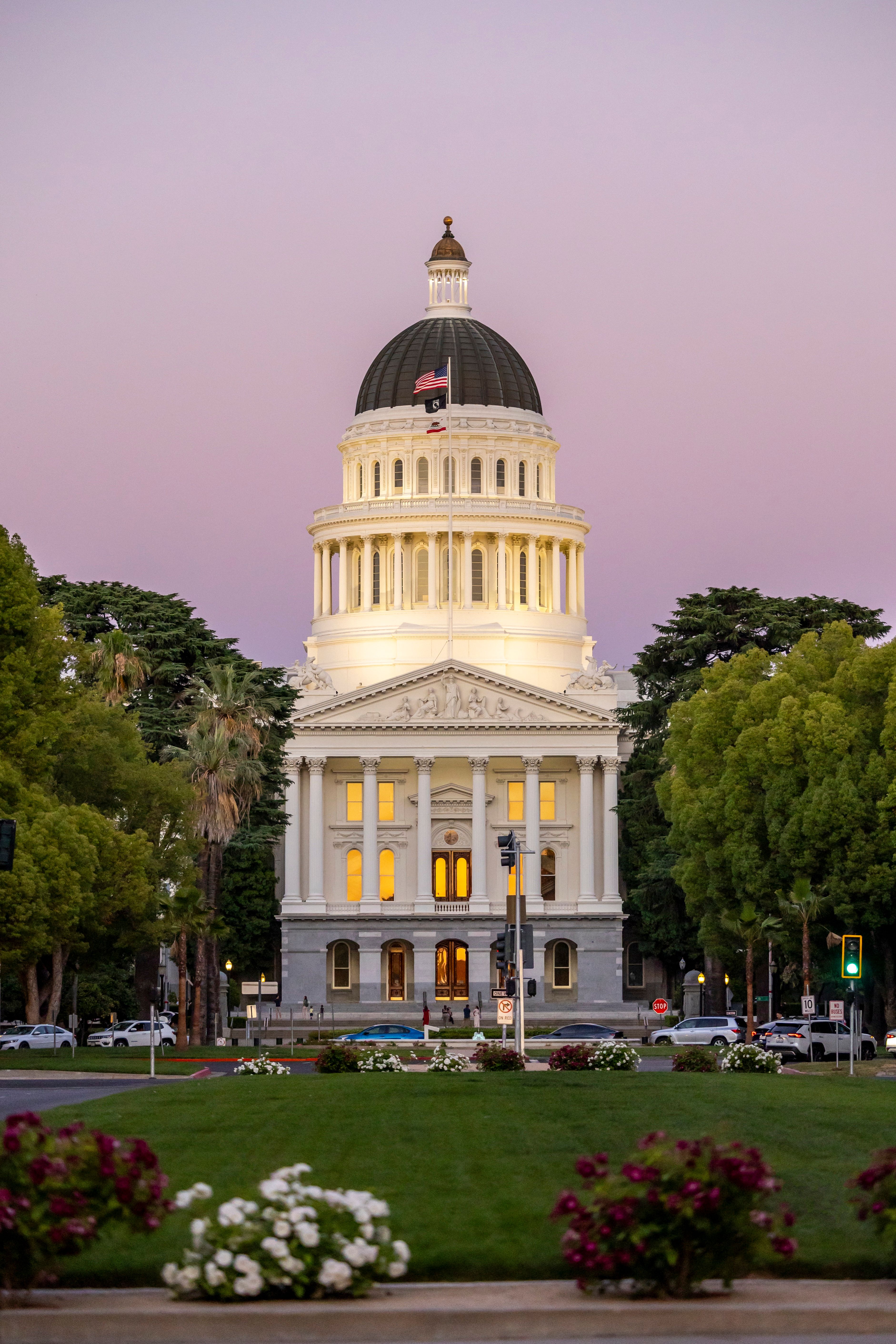 California State Capitol building with dome lit against a purple evening sky, surrounded by trees, flowers, and parked cars, with an American flag flying atop.
