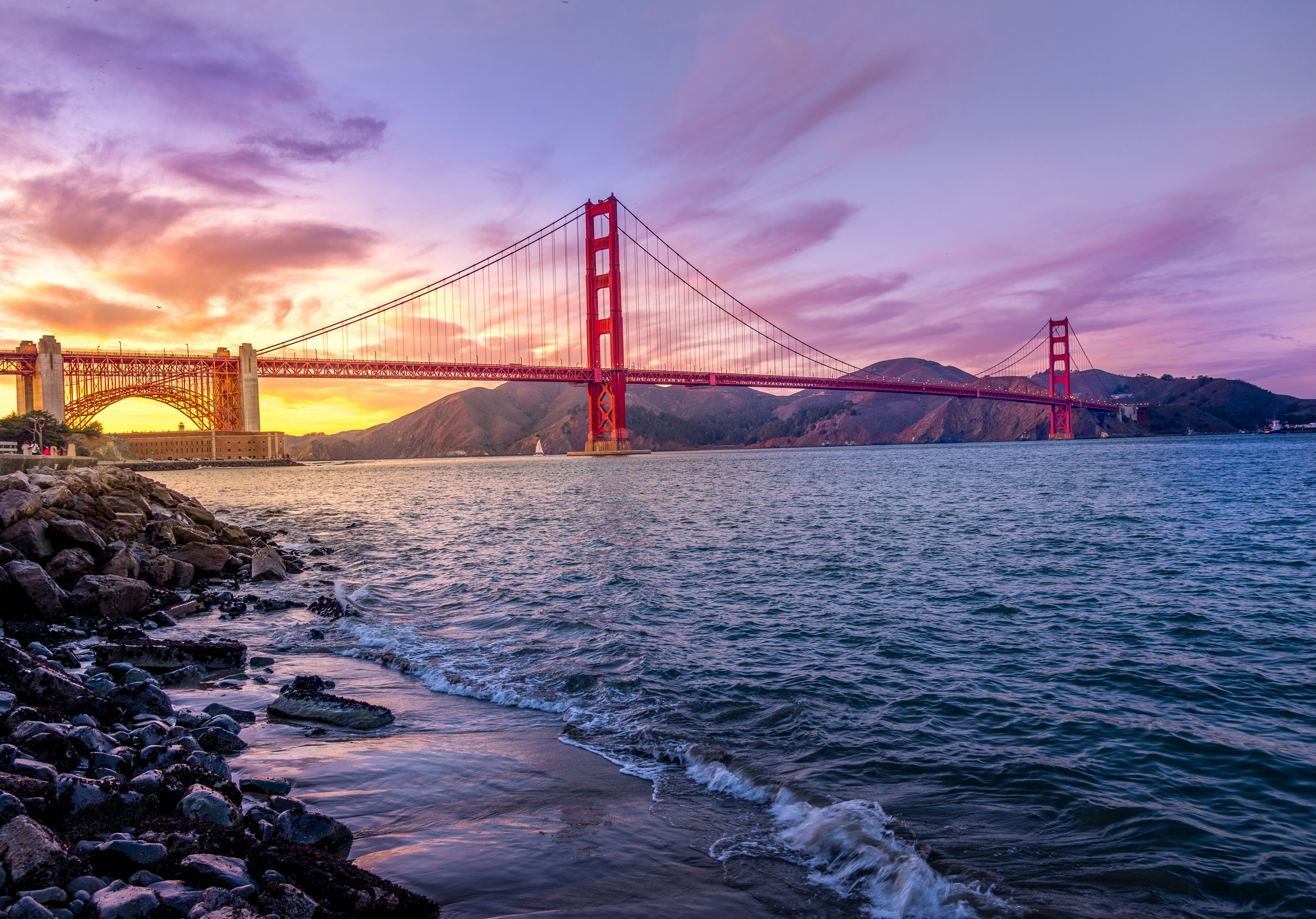 Golden Gate Bridge spanning across a body of water at sunset with rocky shoreline and hills in the background under a colorful sky.