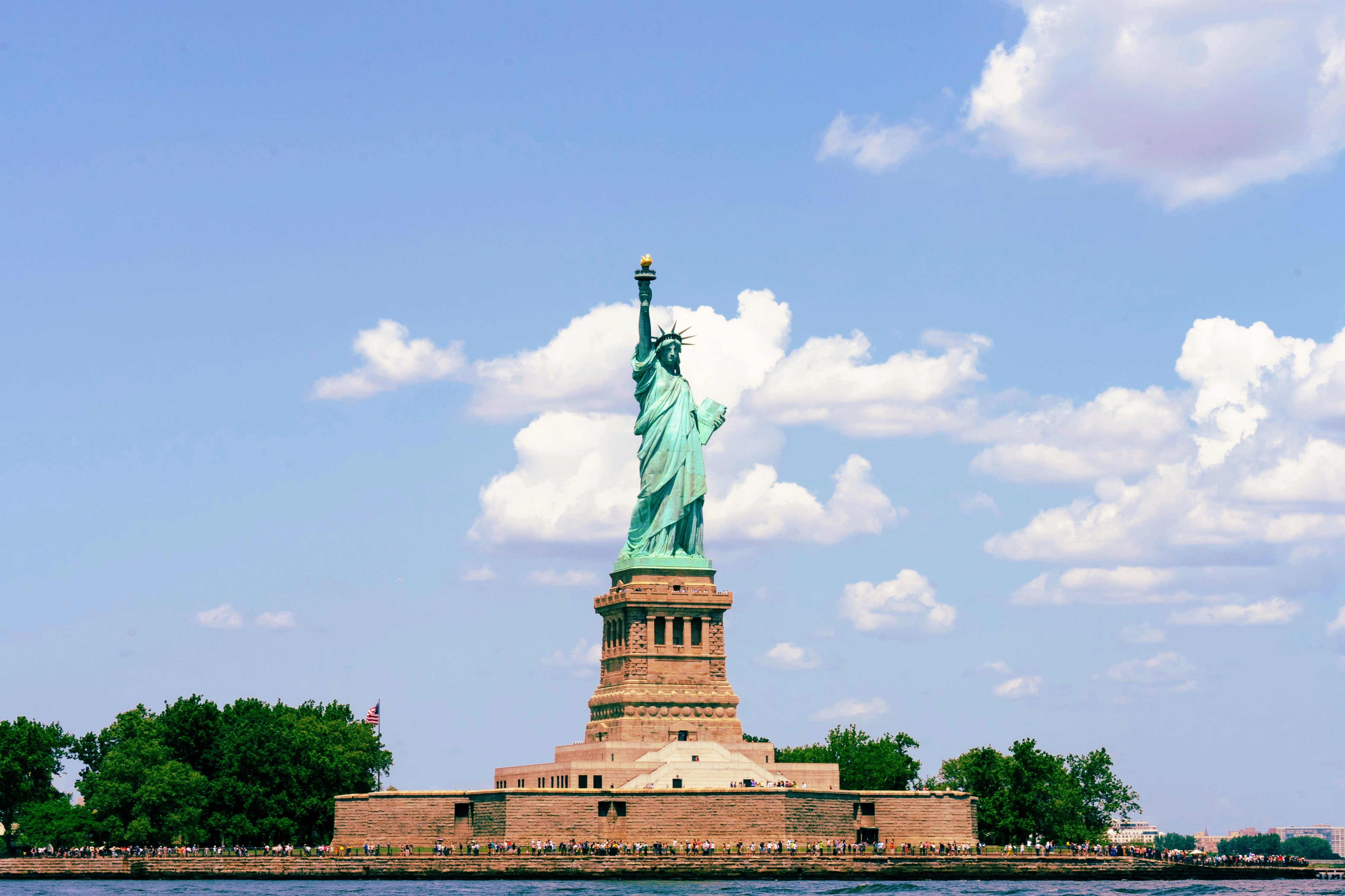 Statue of Liberty on Liberty Island with visitors along the base, under a blue sky with scattered white clouds.