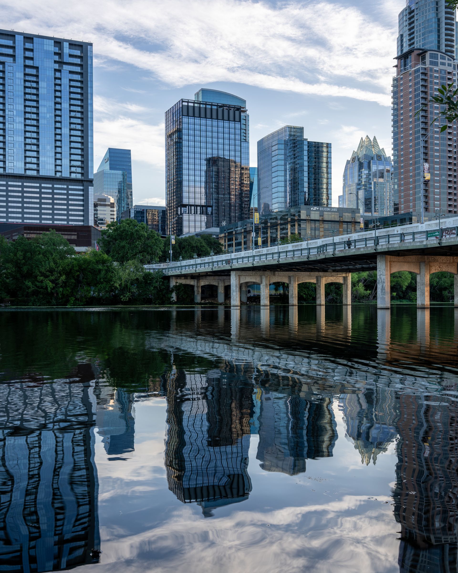 Cityscape of modern glass skyscrapers reflected on the calm river below a partly cloudy sky with a bridge crossing over the water in the foreground and greenery along the riverbank.