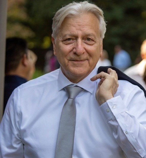 Smiling elderly man with gray hair wearing a white shirt and gray tie, holding a dark jacket over his shoulder outdoors with blurred people and greenery in the background.