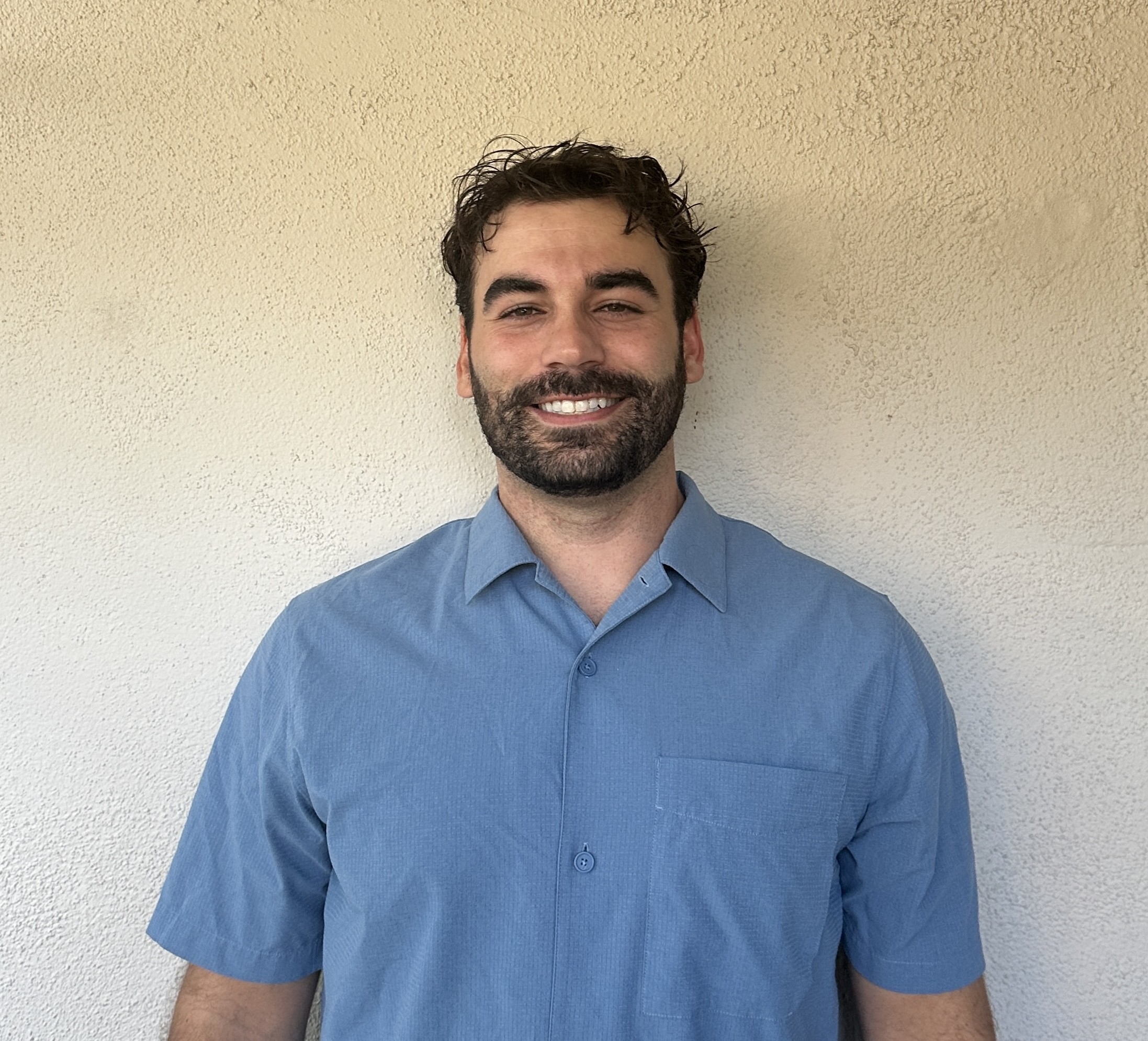 Smiling man with curly dark hair and beard wearing a light blue short-sleeve button-up shirt standing against a textured beige wall.