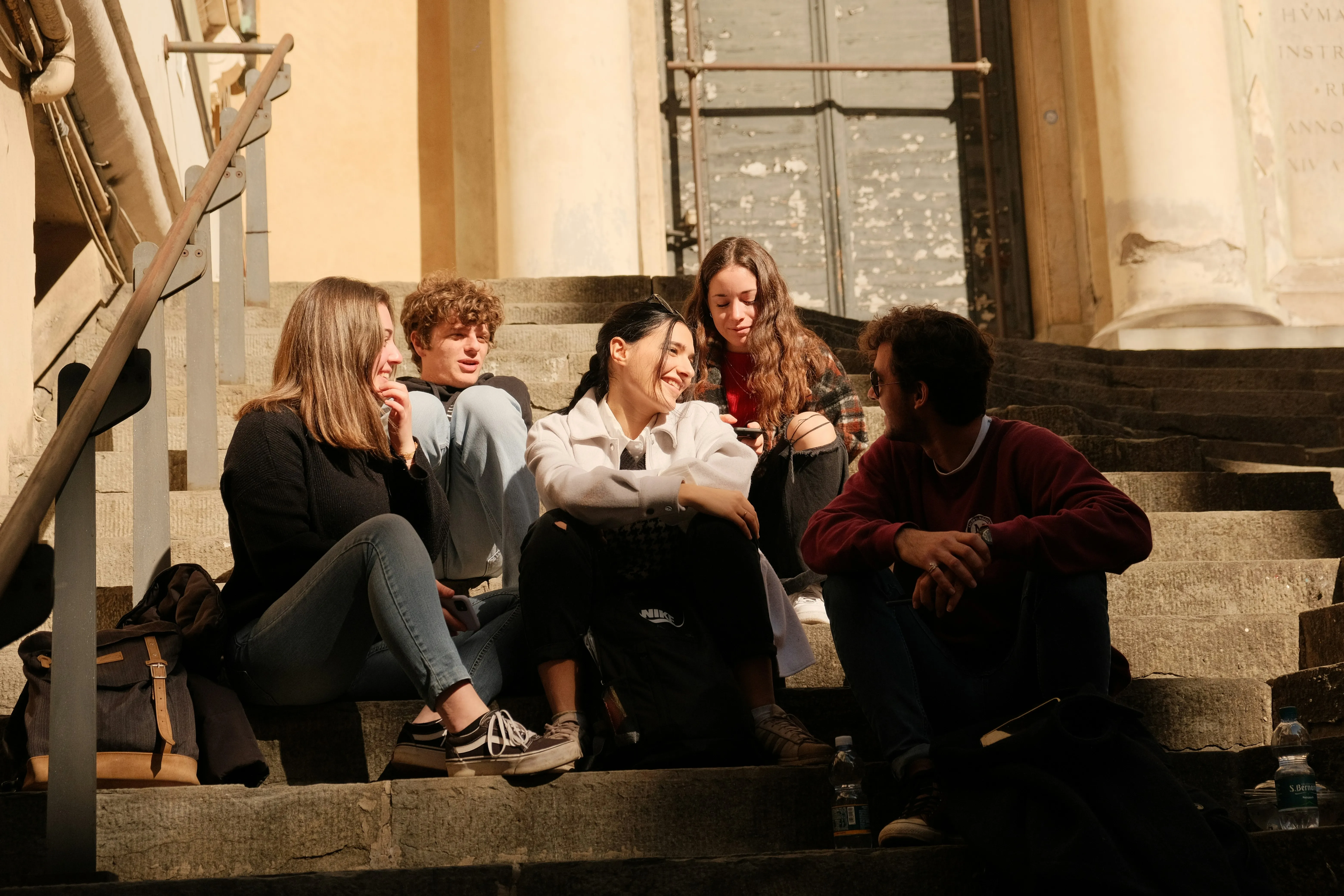 Five young adults sitting on sunlit stone steps, chatting and smiling in a relaxed outdoor setting.