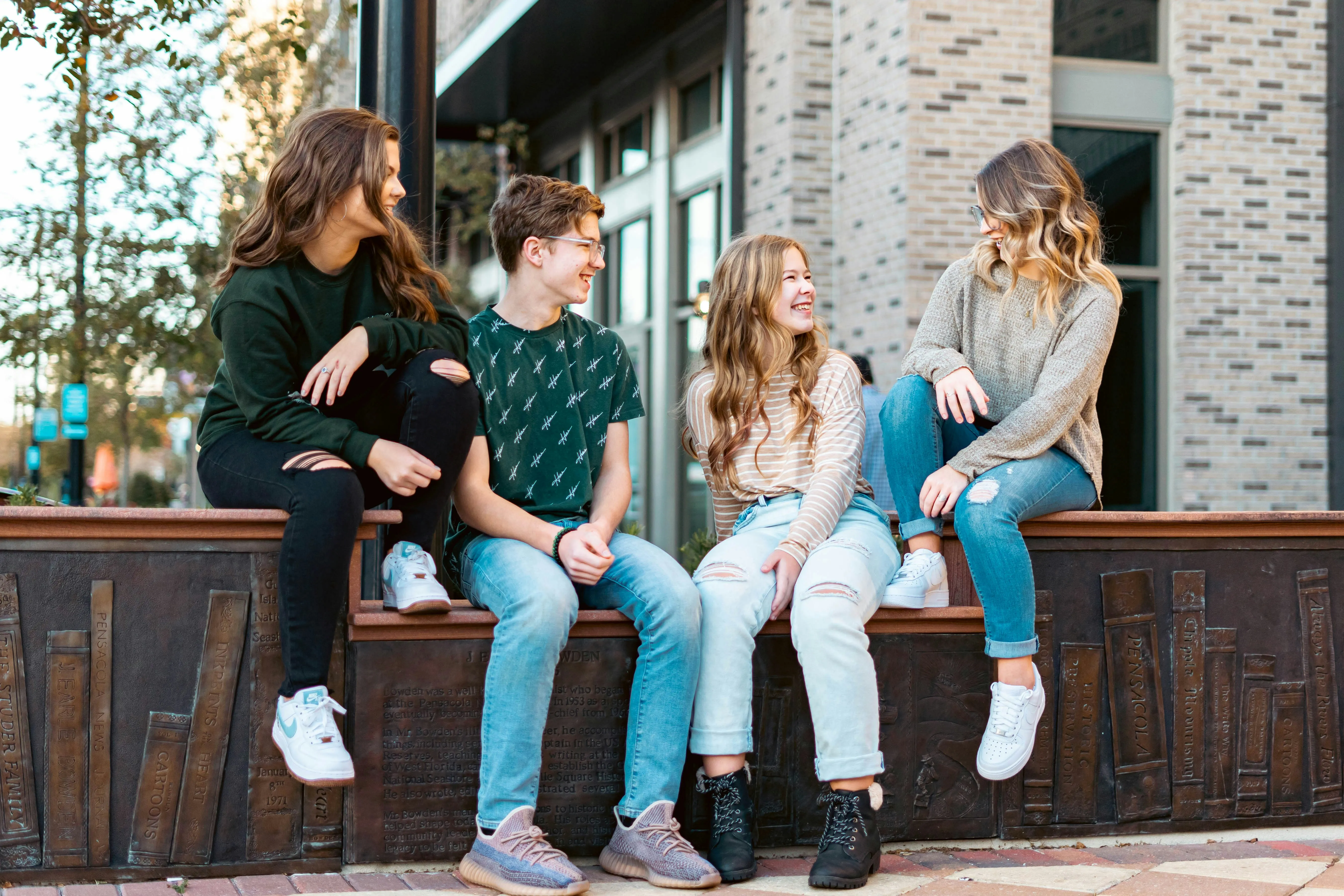 Four young friends sitting on a low wall outside, smiling and talking together in casual clothes.