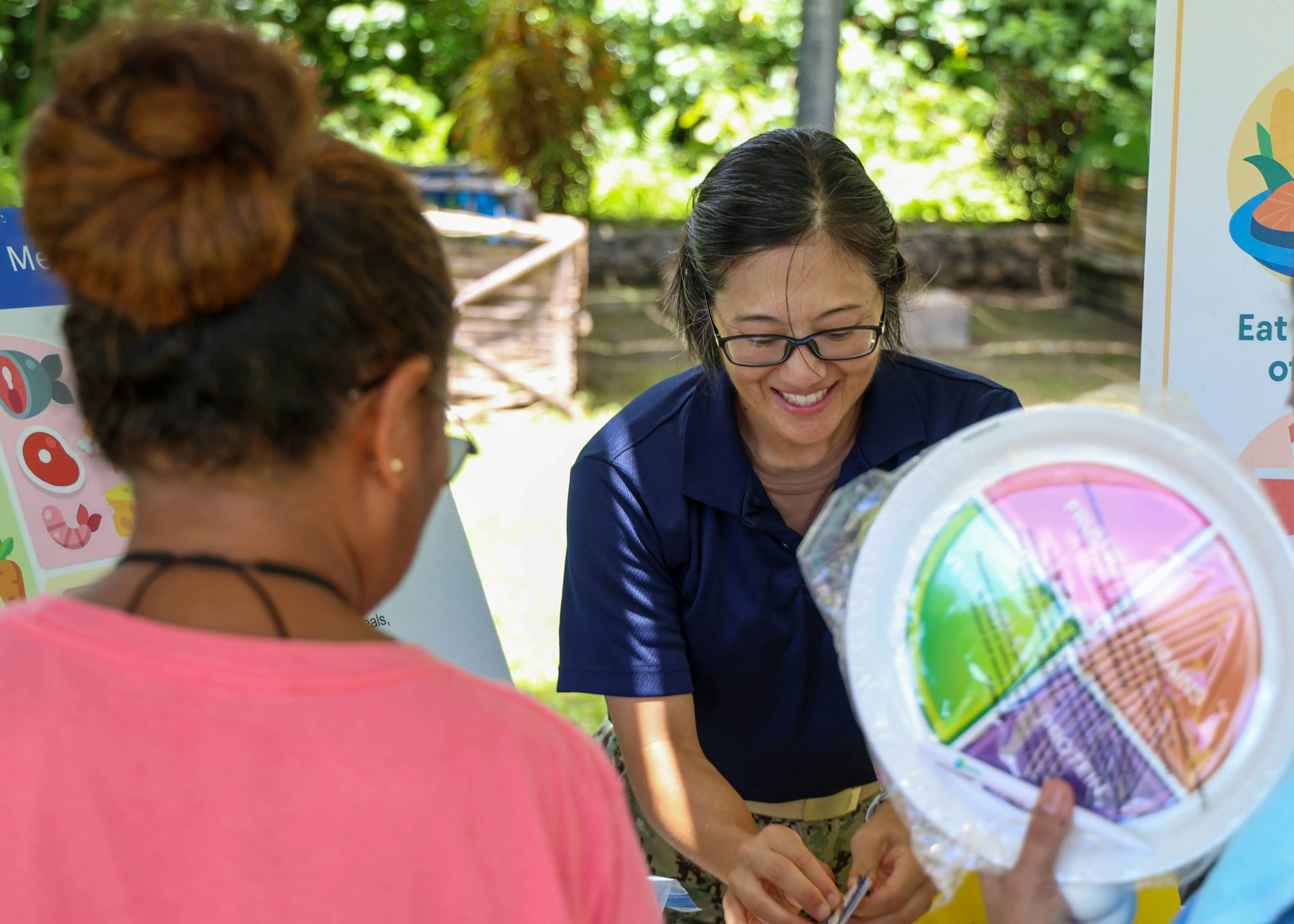 Woman in navy shirt smiling and interacting with a person holding a colorful nutrition plate outdoors.